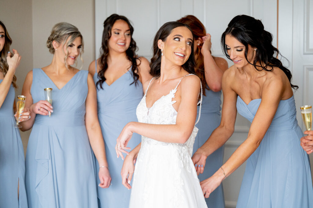 a bridesmaid in a pale blue dress helps zip up the bride's wedding gown