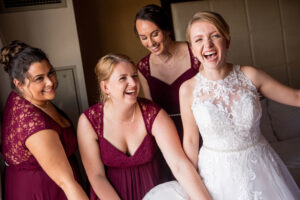 Bride laughing with your bridesmaids as she gets in your wedding dress 