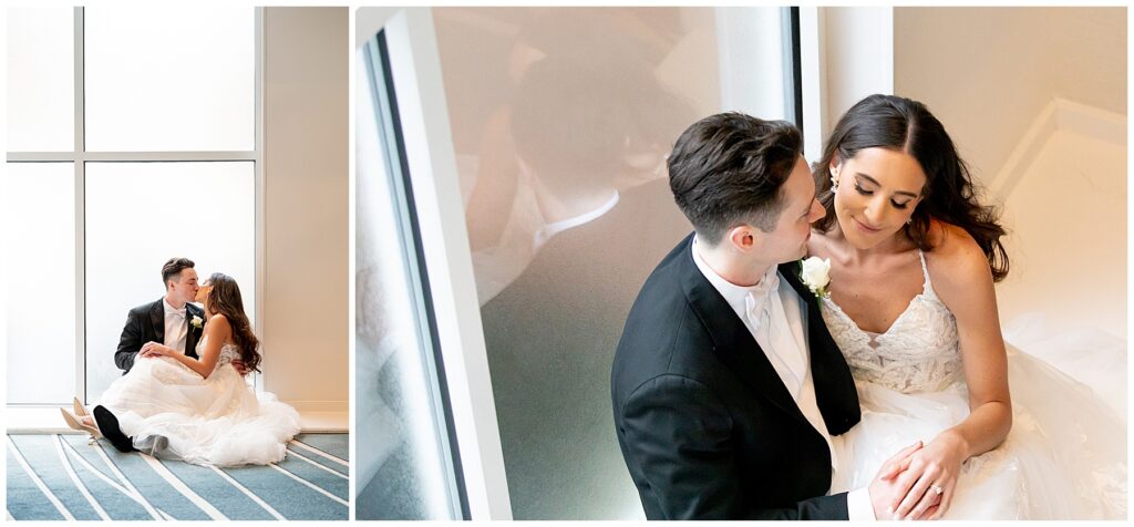 bride and groom sit on the floor by the large windows and kiss