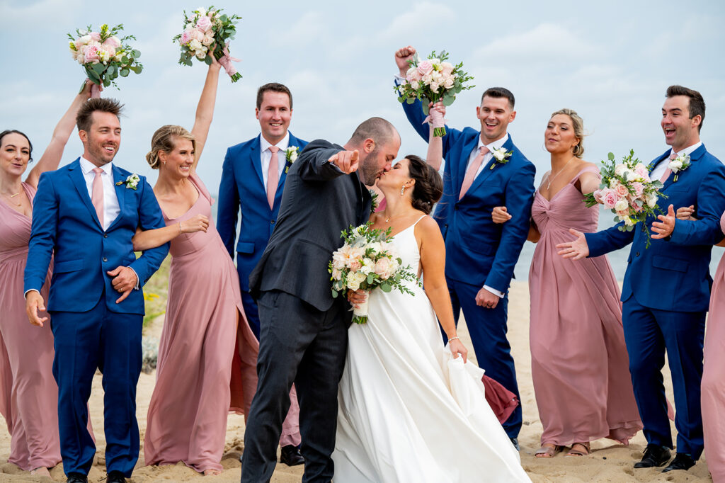 bride and groom kiss as wedding party in blue suits and pink dresses celebrate behind them