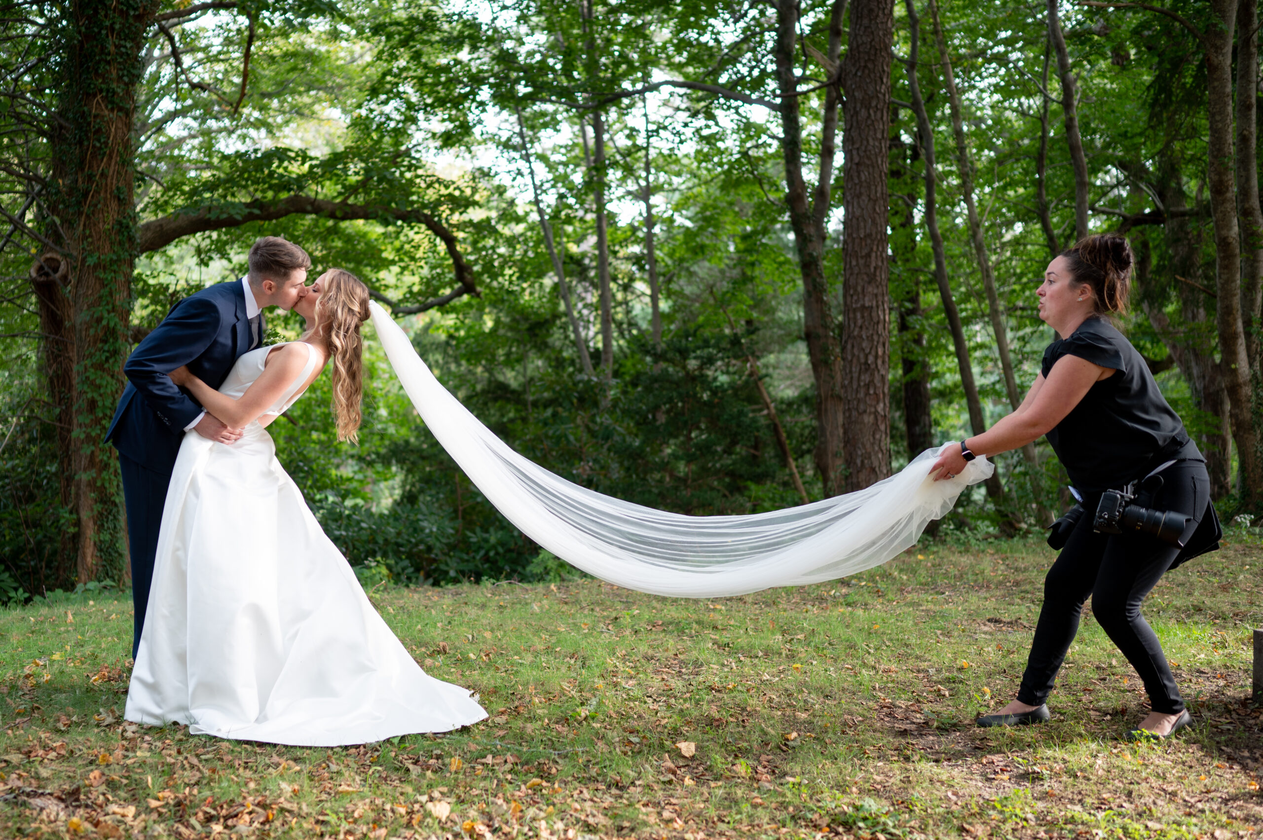 bride and groom kiss in the woods while second shooter holds veil 