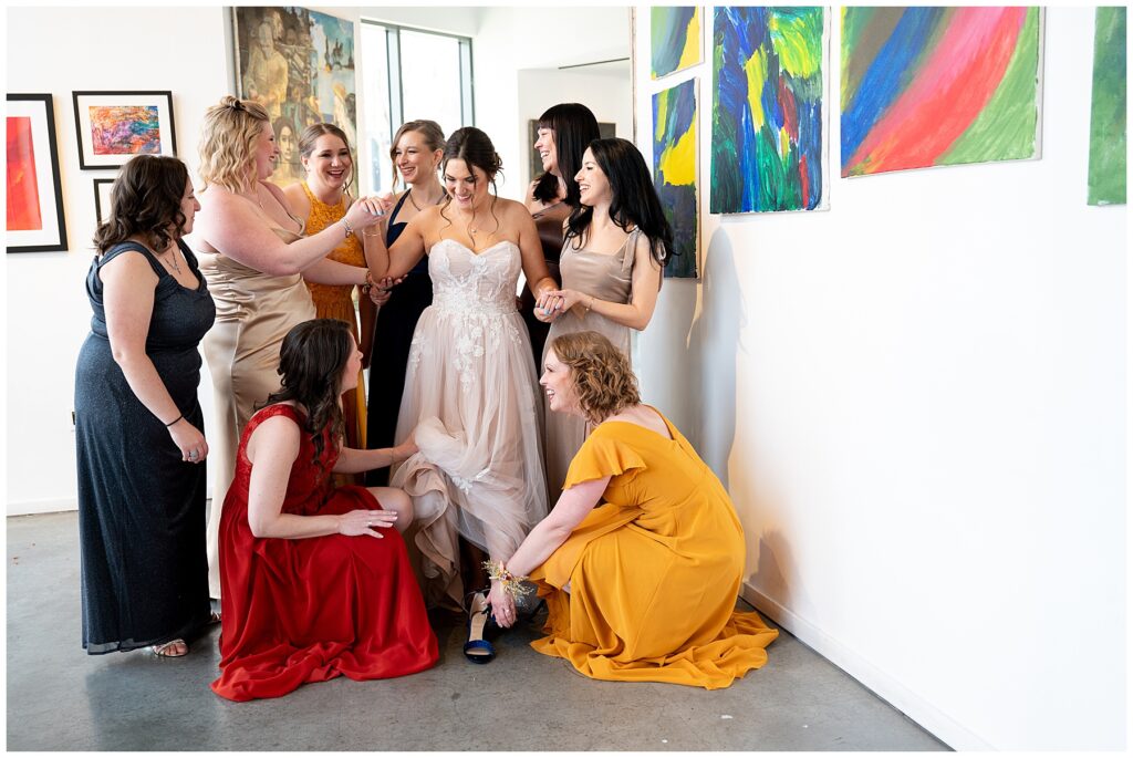 bridesmaids in black and yellow and red dresses help the bride put on her shoes