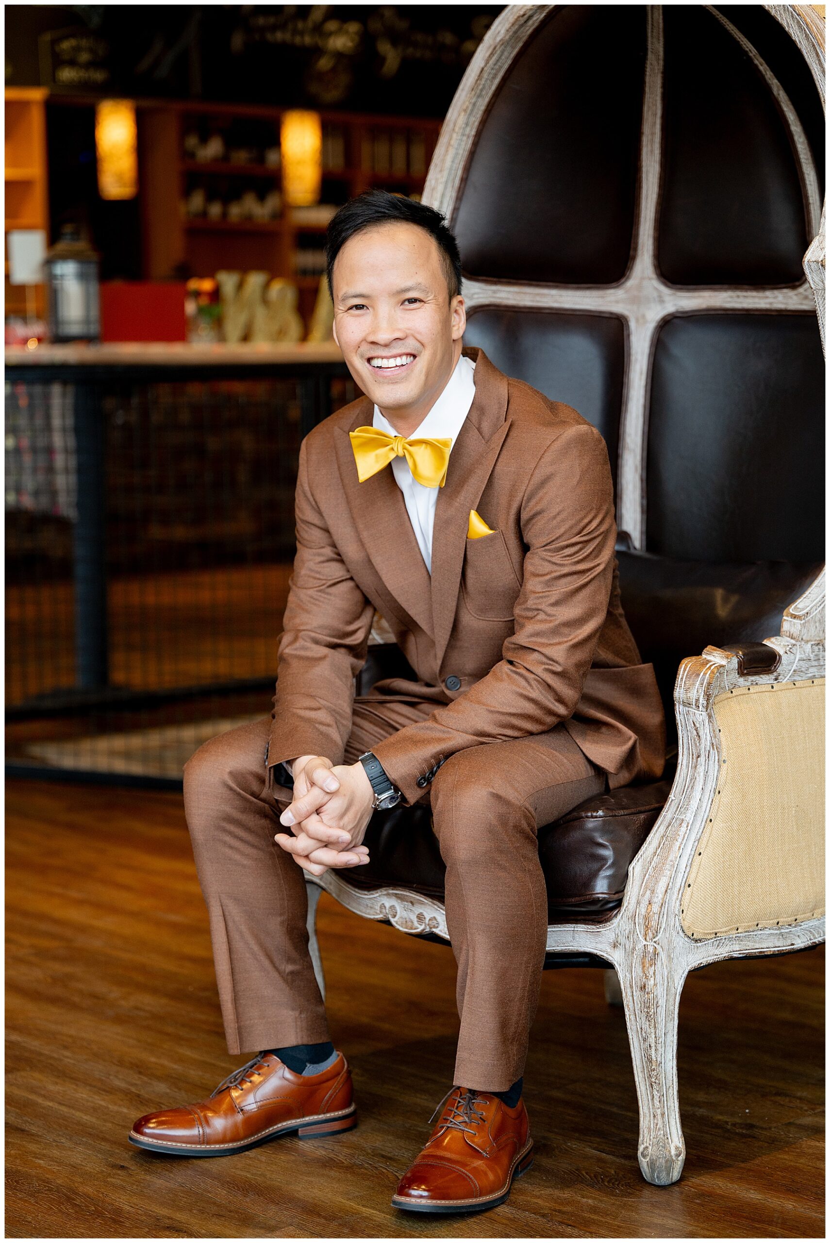 groom sits in chair wearing brown suit and yellow bowtie smiling at the camera