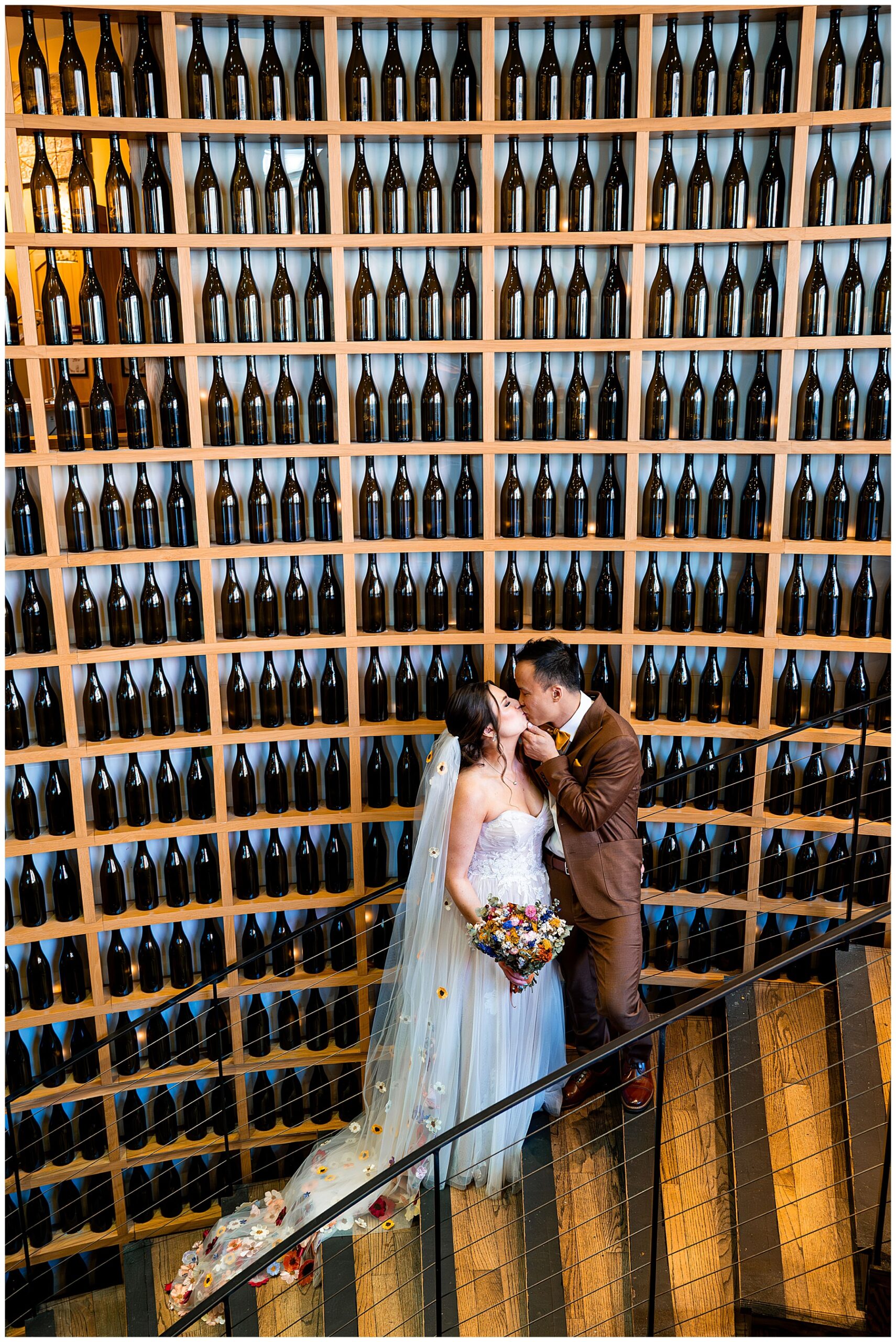 bride and groom kissing on the stairs in front of wall of wine bottles