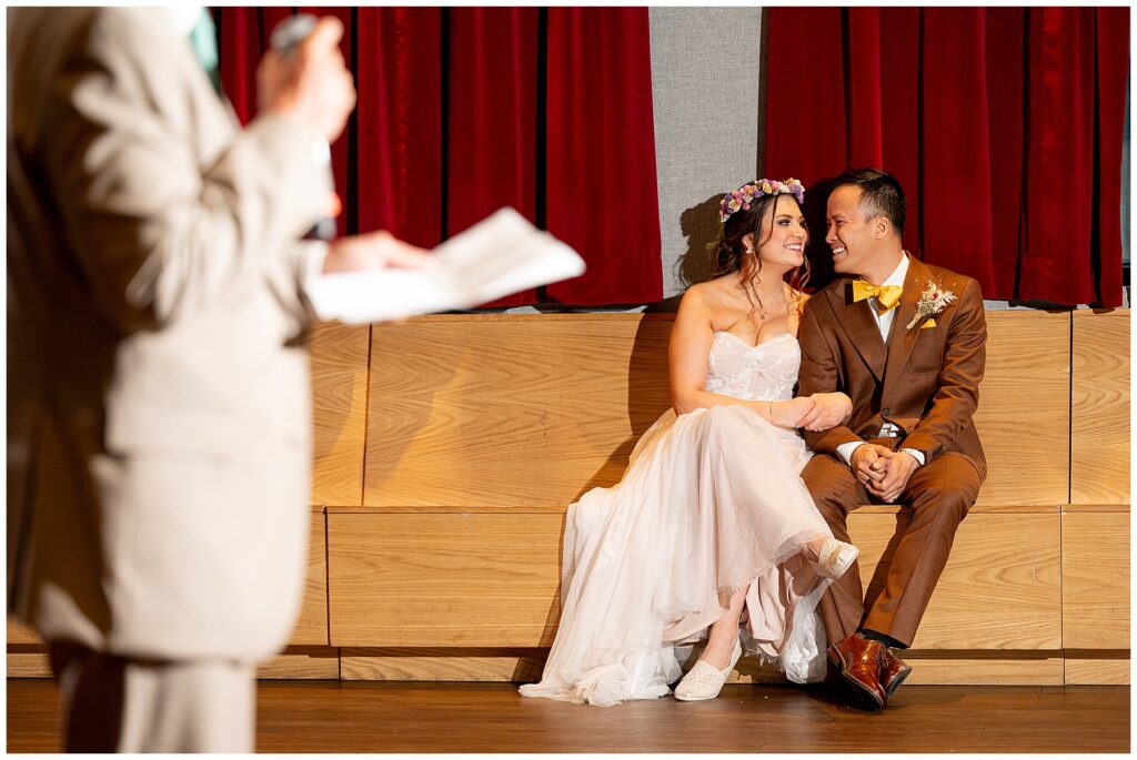 bride and groom smile at each other while father gives a toast 