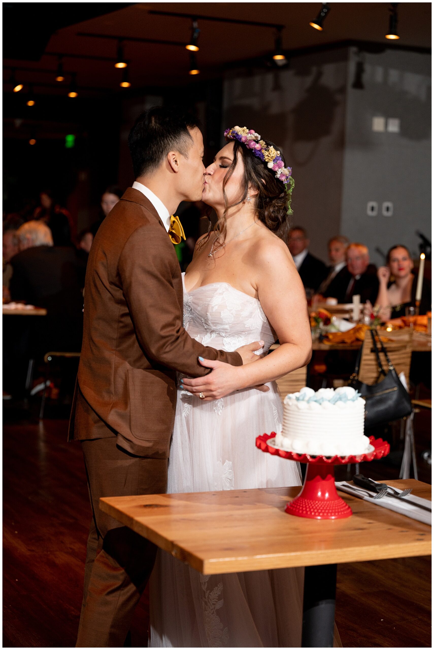 bride and groom kiss after cutting the cake as guests watch