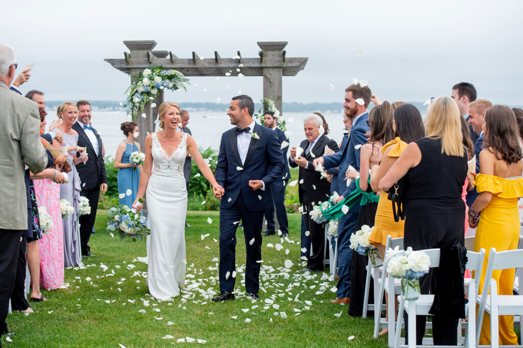 bride and groom holding hands and smiling as they walk down the aisle as guests throw flower petals