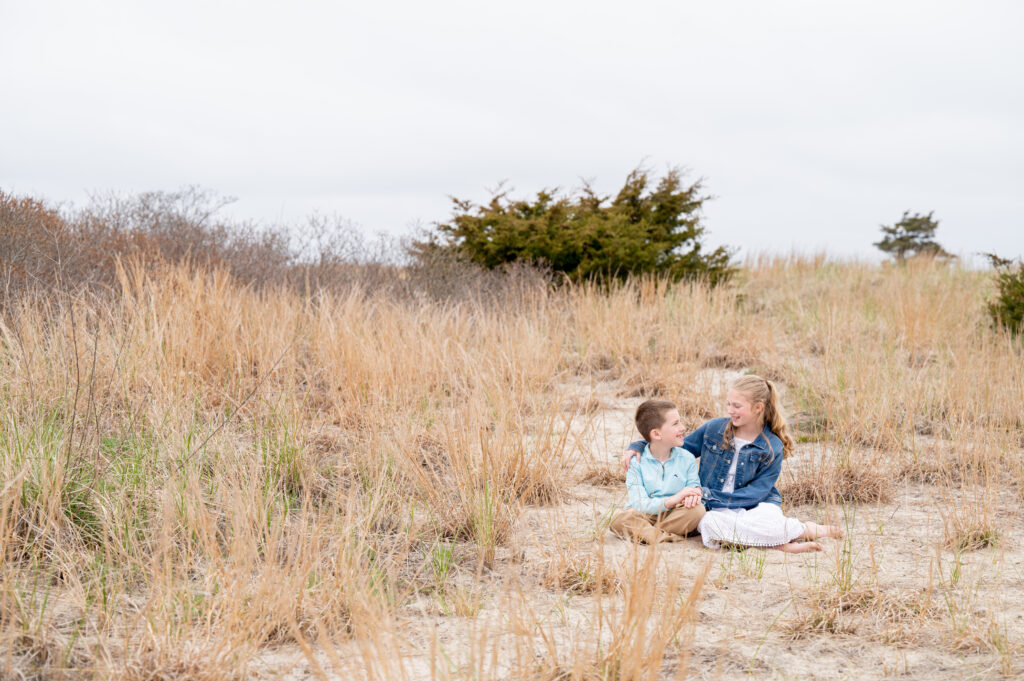 a brother and sister sit on the beach smiling at eachother 