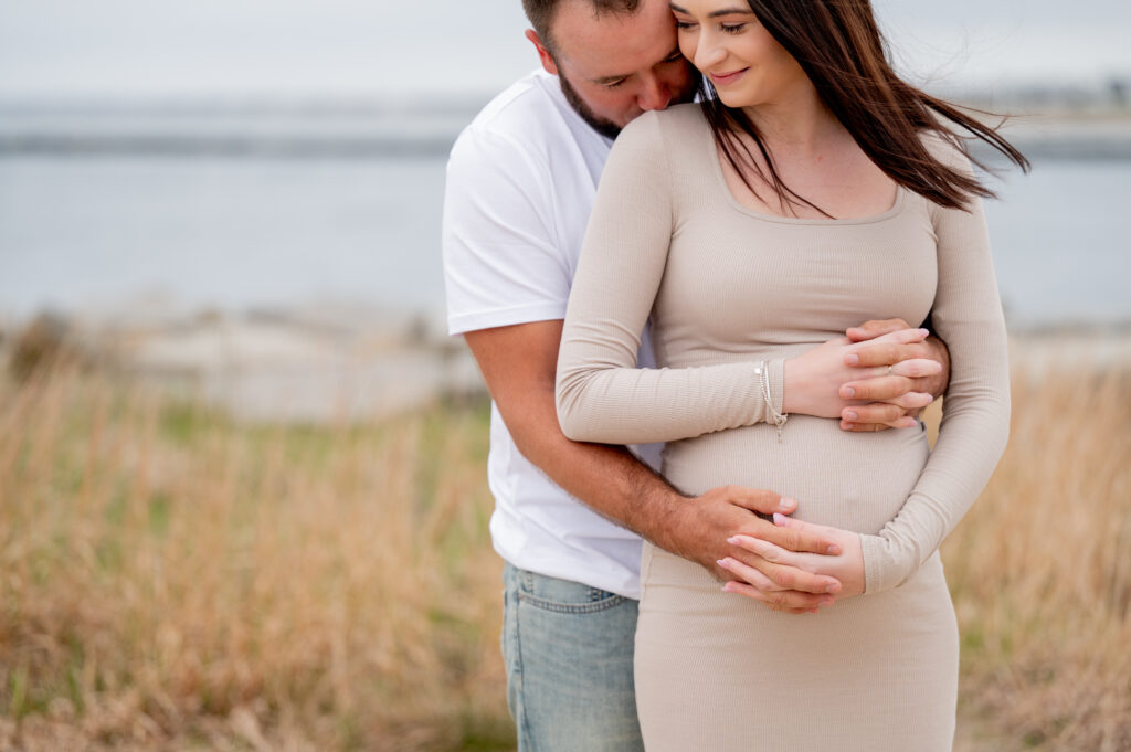 a husband hugs his pregnant wife from behind while she smiles and holds his hand wearing a tan dress on the beach 