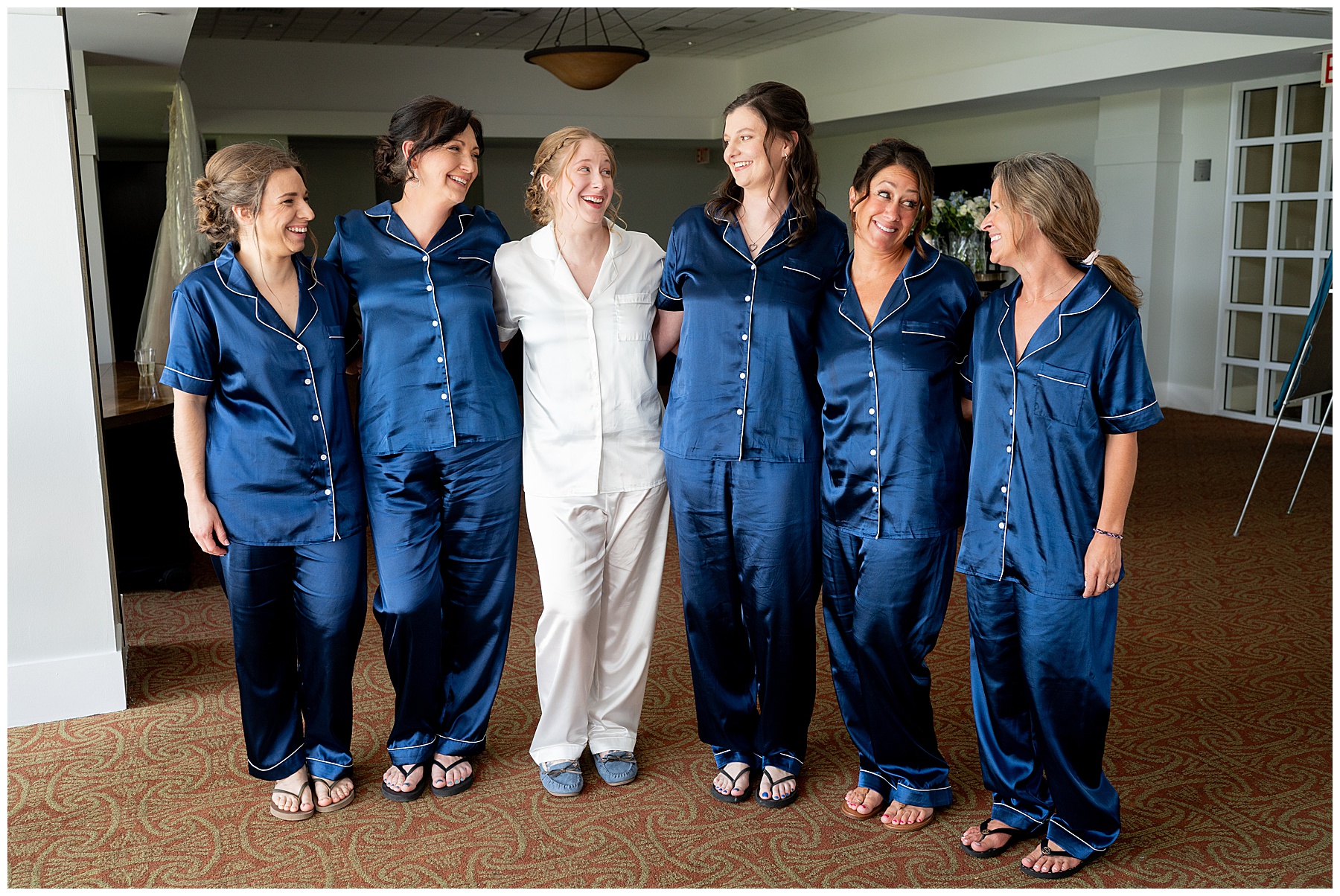 the bride in white pajamas stands with her bridesmaids as they wear dark blue satin pajamas on the wedding day