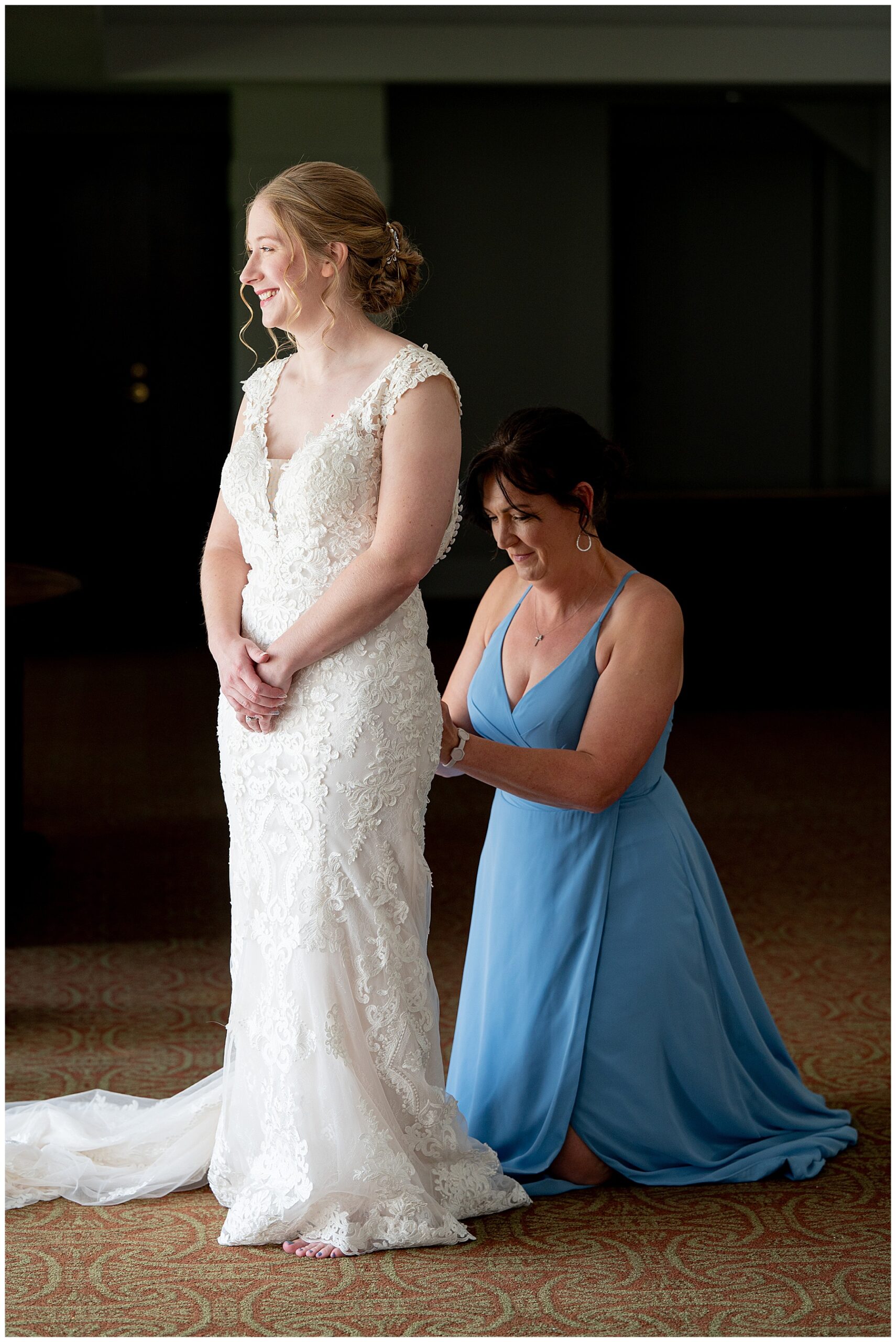 bride kneels down in a light blue dress behind the bride to button up her white wedding dress 