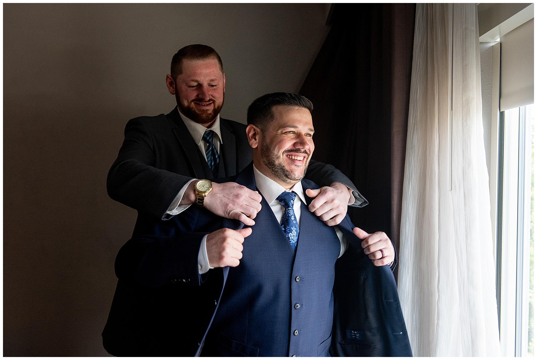 a groomsmen stands behind the groom to help him put his blue suit jacket on 