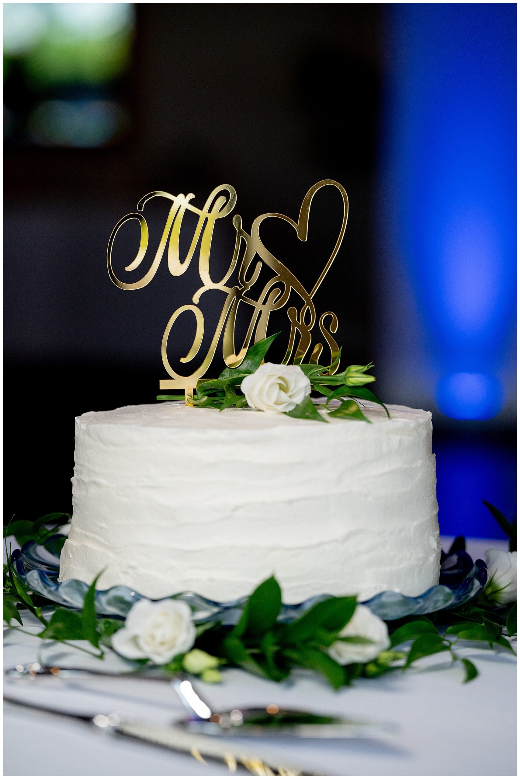 a white wedding cake with green flowers and a gold mr & mrs cake topper made by Milk & Honey Bakery a Leominster, Massachusetts based bakery