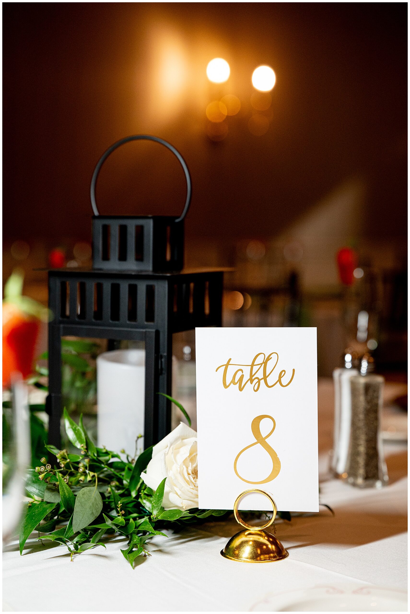 table centerpiece with a black lantern and greenery with a table number of gold font on a white card