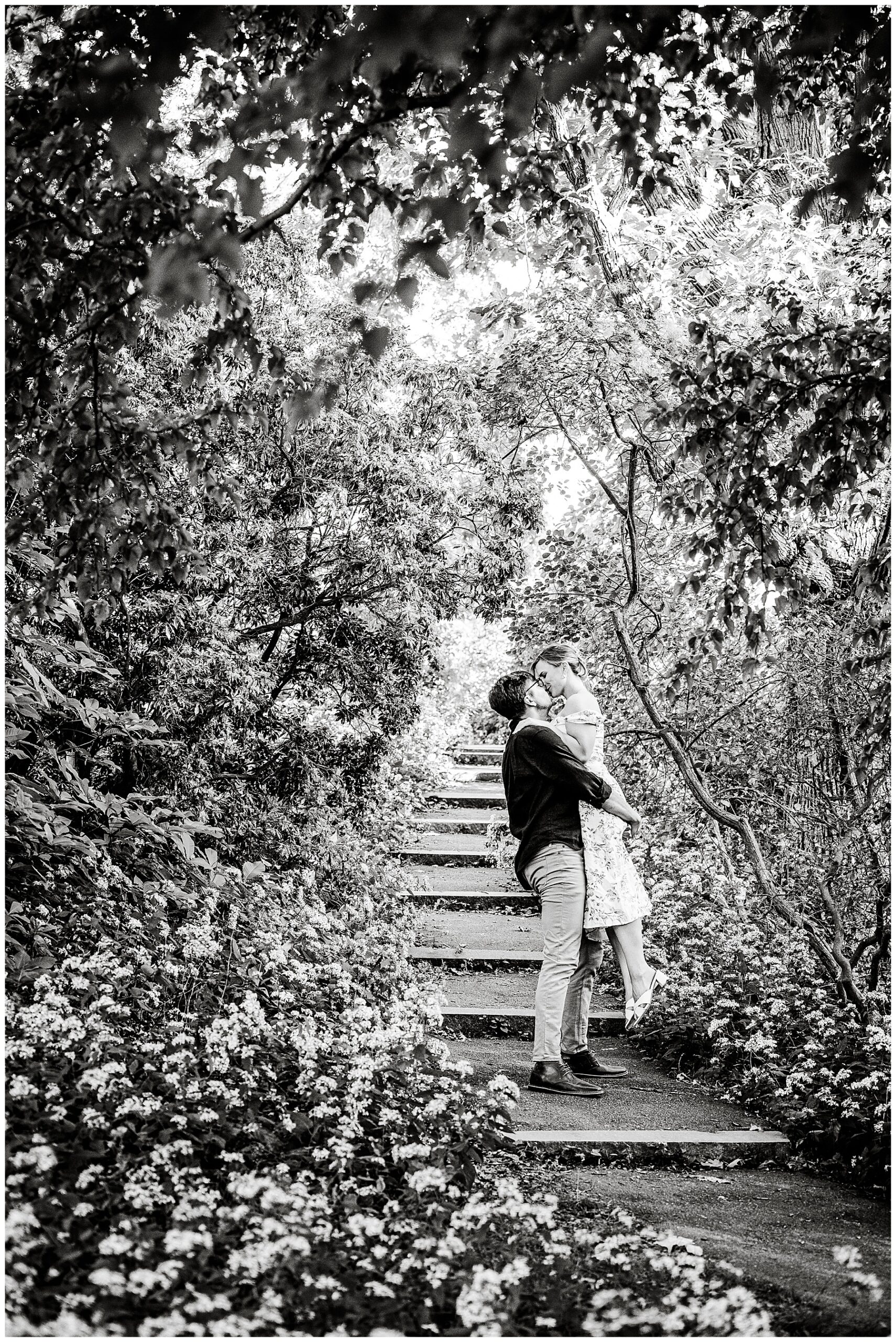 black and white photo of man picking up and holding his fiance as they kiss on the steps 