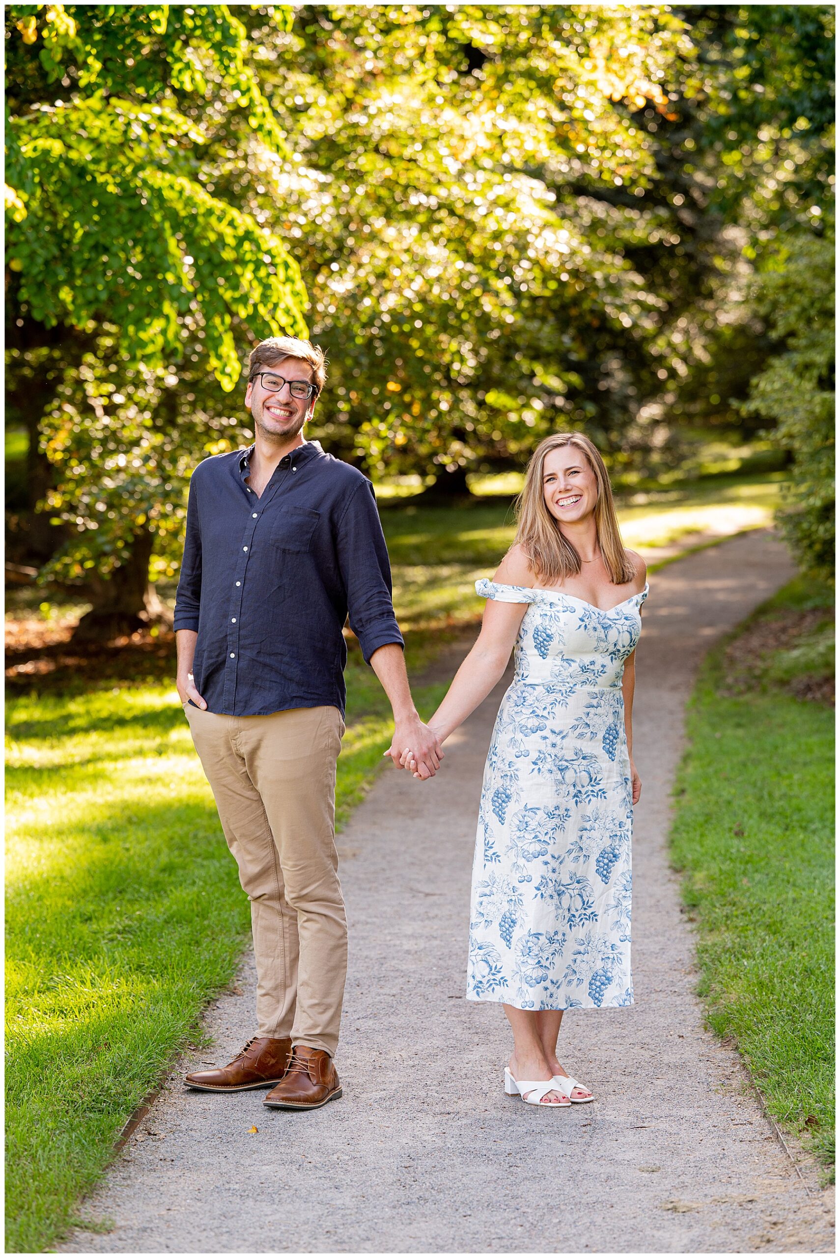 man in khakis and blue button down shirt and woman in white and blue patterned dress stand holding hands smiling at the camera