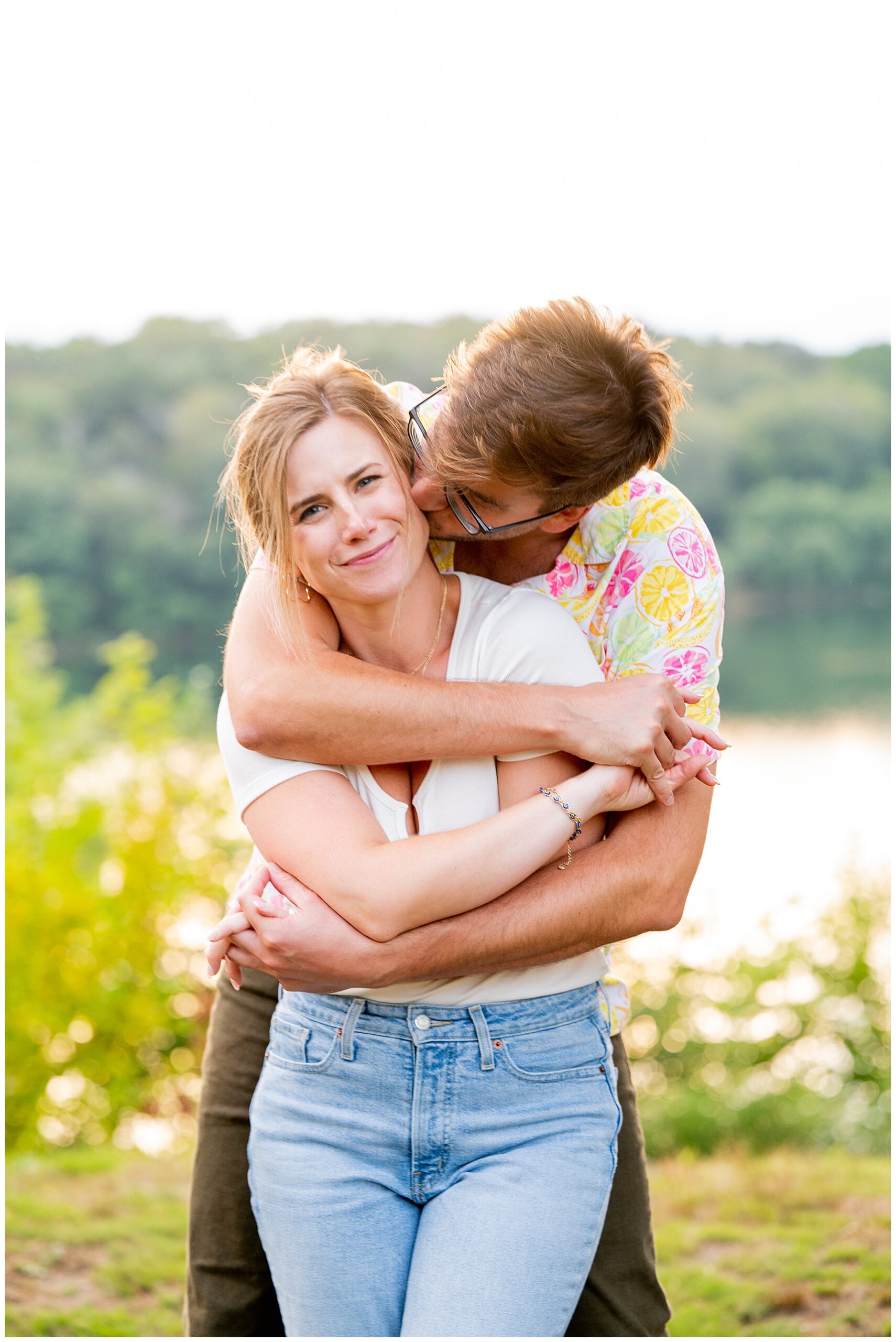 man hugging his fiance from behind and kissing her cheek as she smiles at the camera 