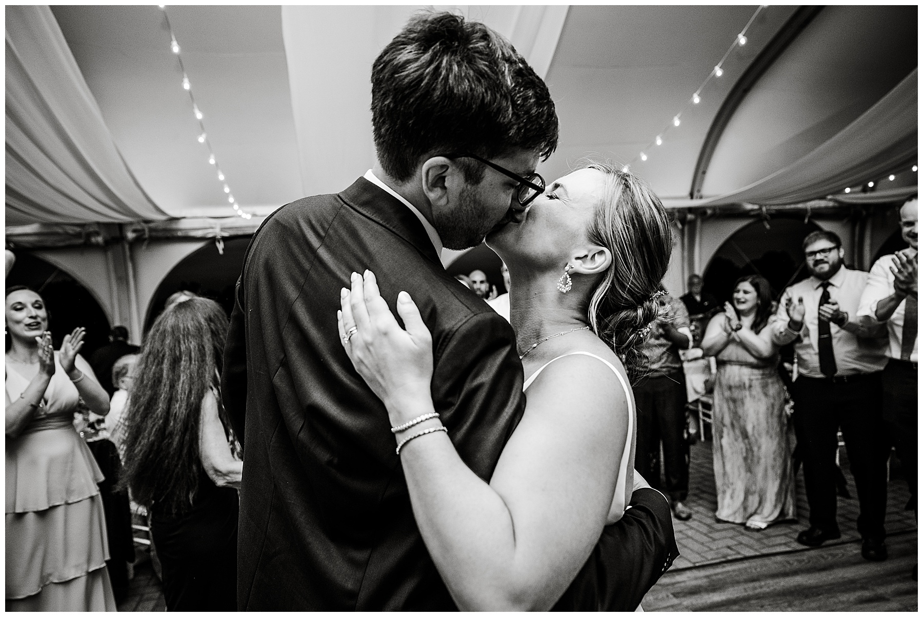 black and white photo of bride and groom kissing on the dance floor surrounded by guests 