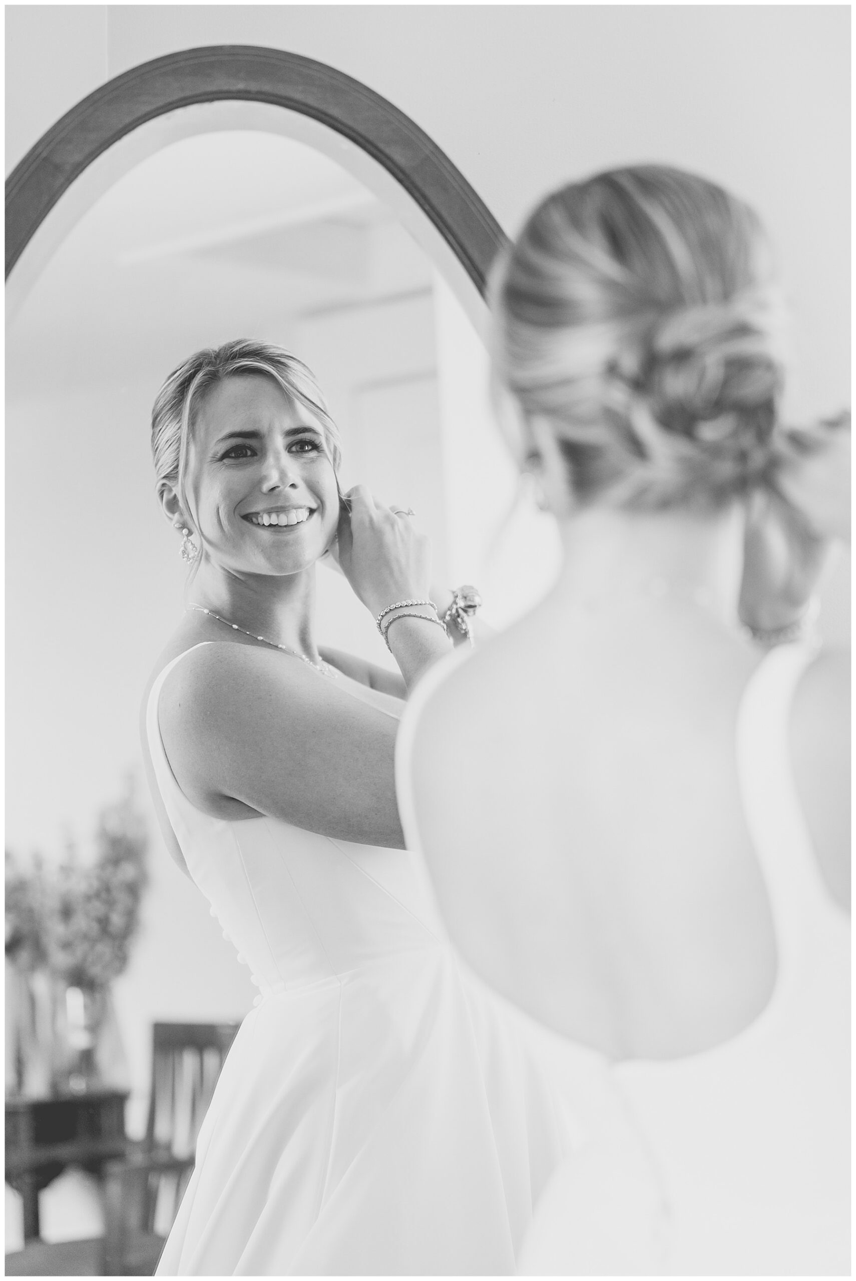 black and white photo of bride smiling into the mirror as she puts on her wedding earrings 