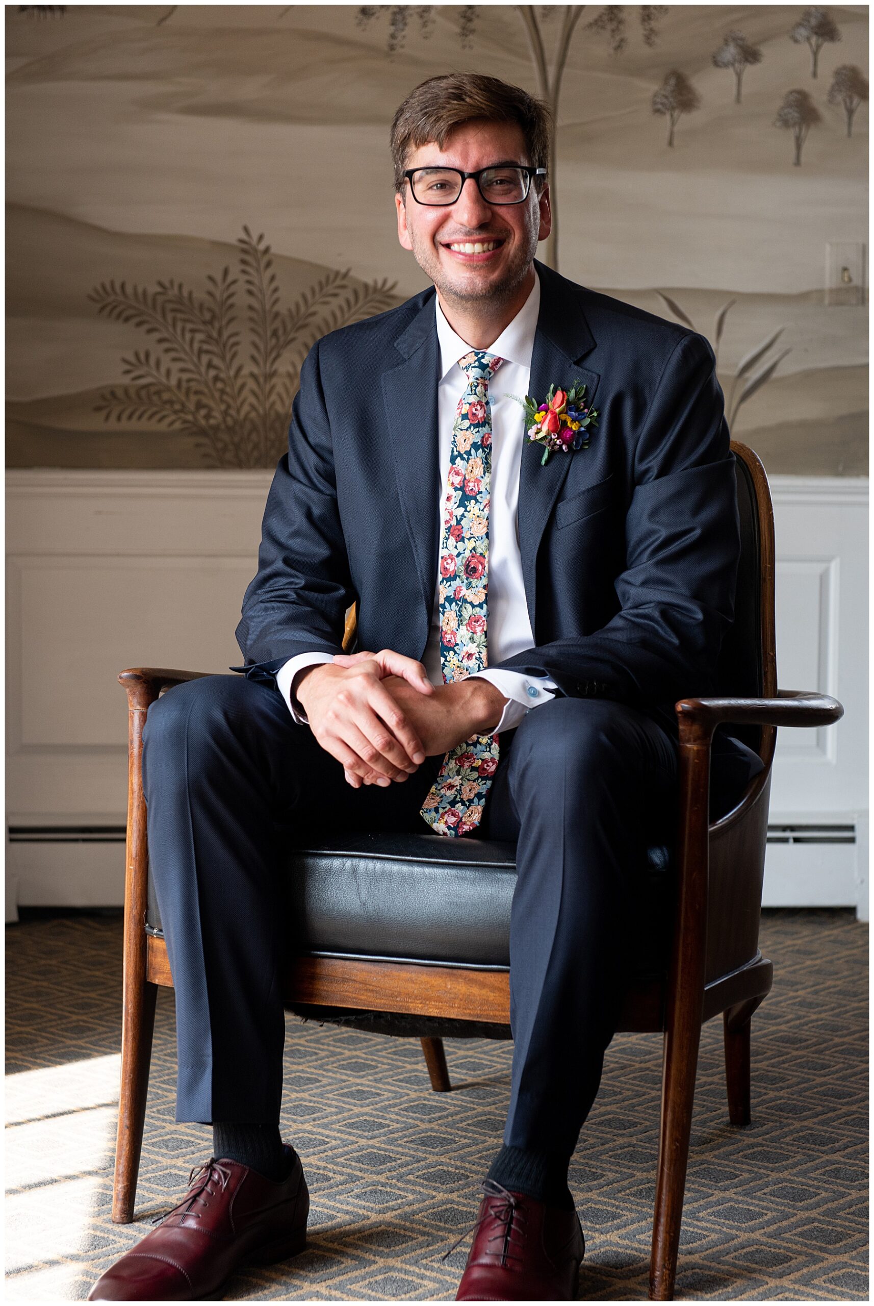 groom sitting down and smiling at the camera wearing a blue suit and floral patterned tie 