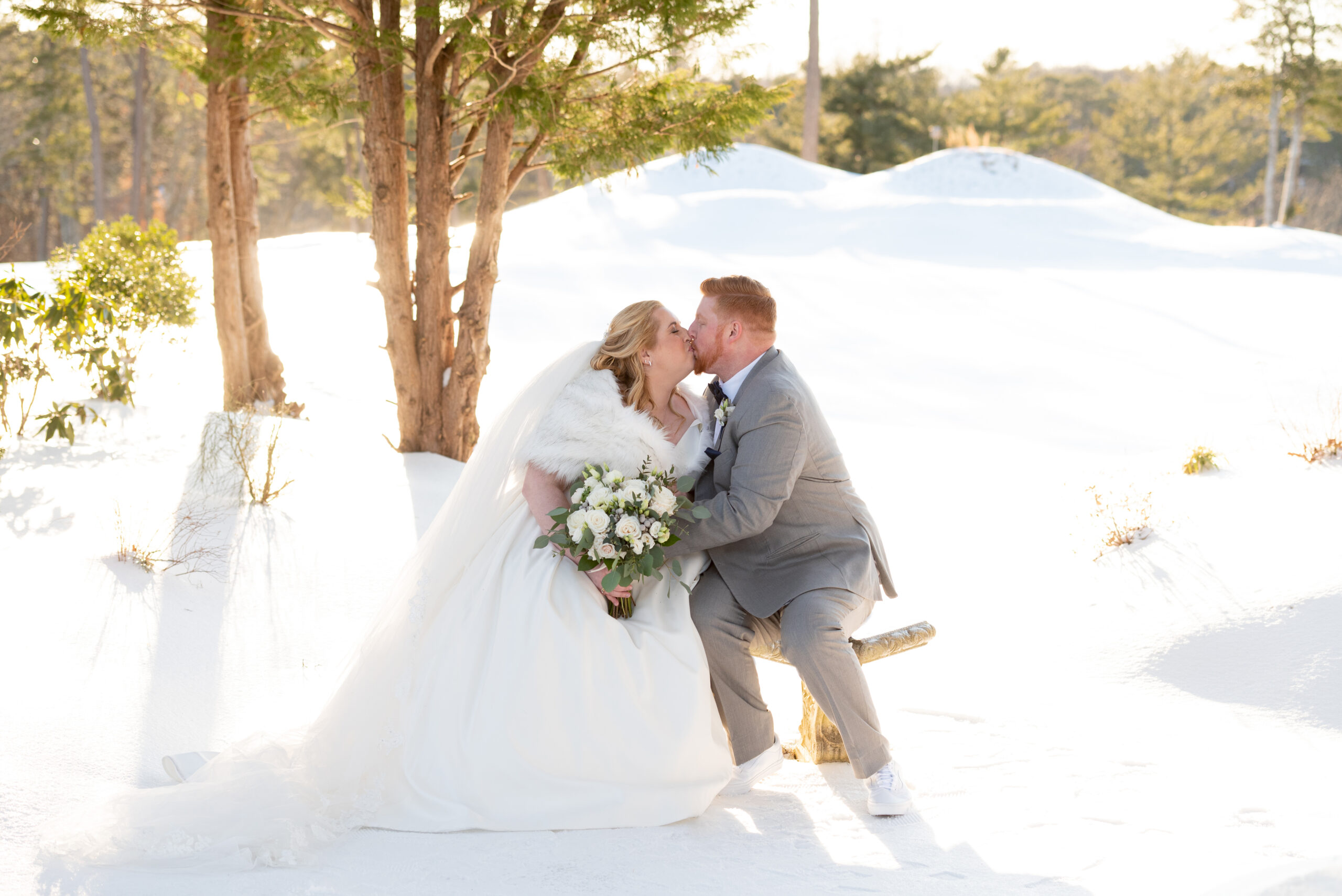 bride and groom sitting on a bench outside kissing surrounded by snow