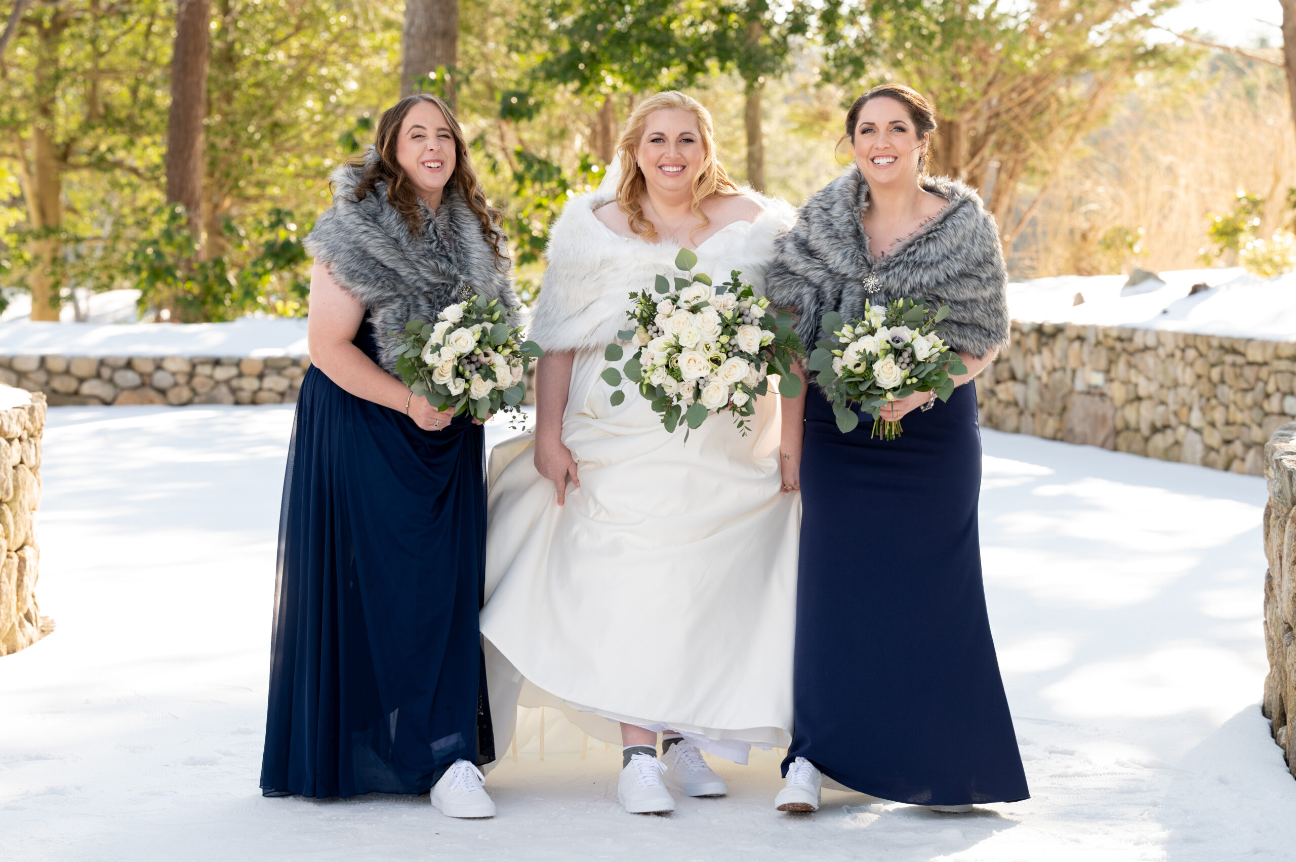 bride wearing her wedding dress and white shawl standing outside with her bridemaids wearing navy blue dresses and grey shawls with a snowy background