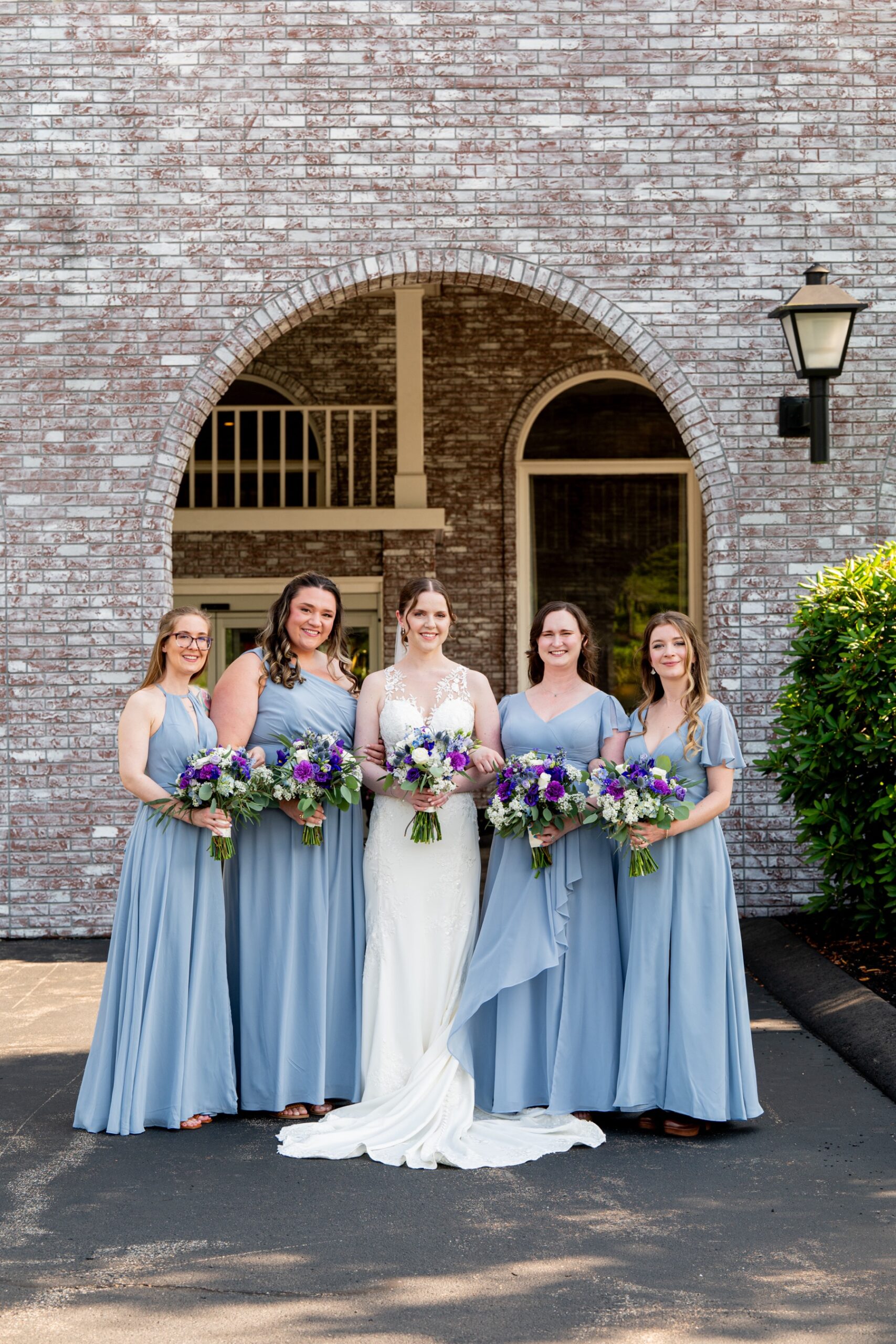 bride standing with her bridesmaids who wear light blue dresses and holding purple and white colored bouquets 