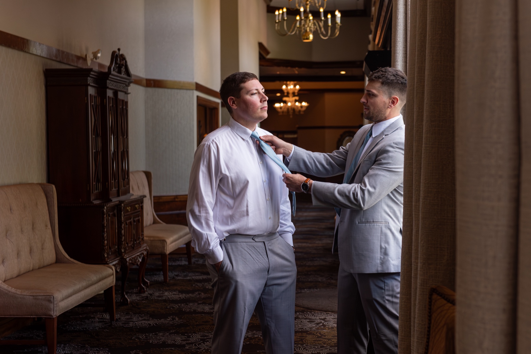 groomsman wearing grey tux helps groom out his tie on