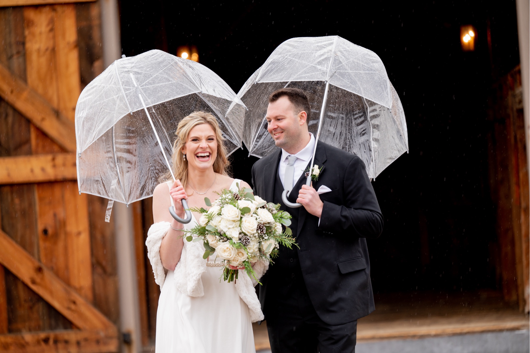 bride and groom smiling and laughing as they walk carrying clear umbrellas 