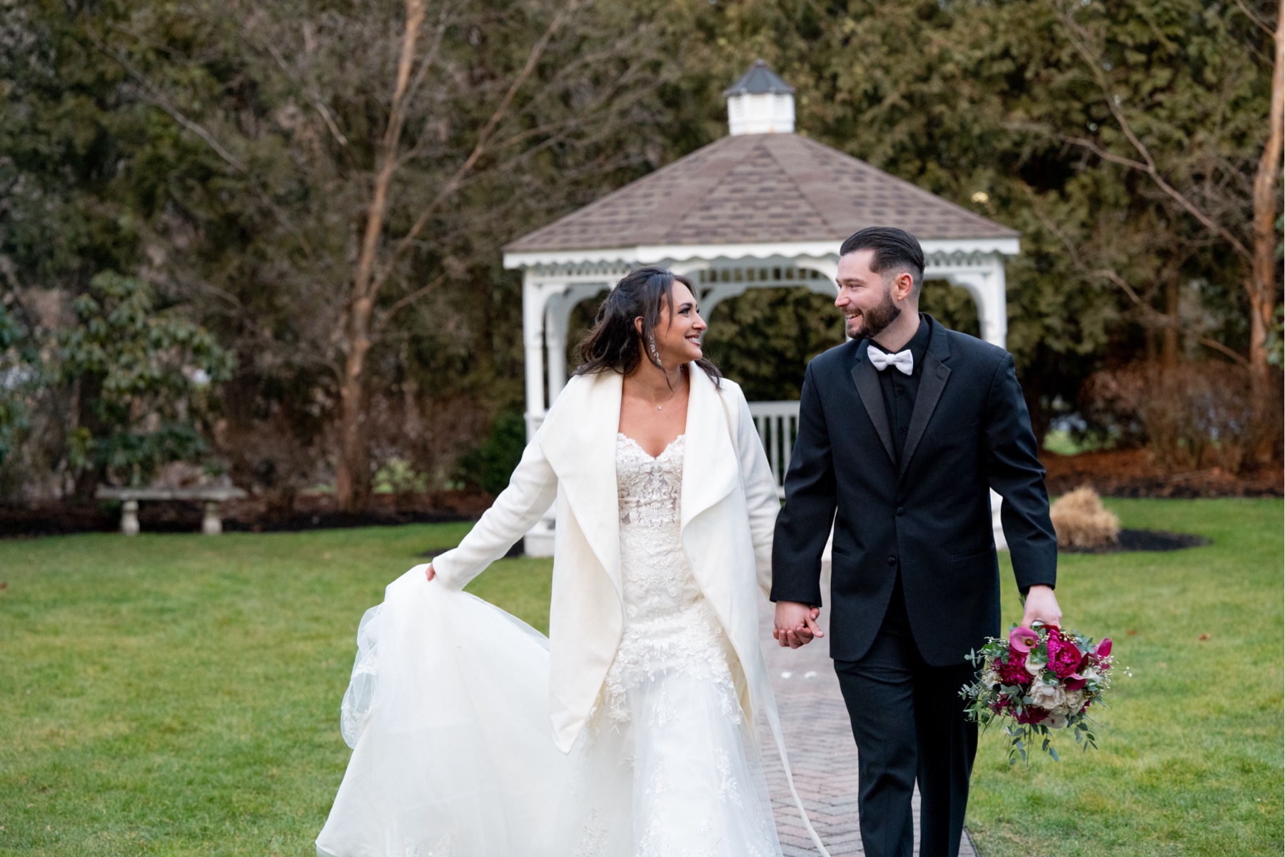 bride walks along wearing white coat with the groom who holds her pink and green wedding bouquet