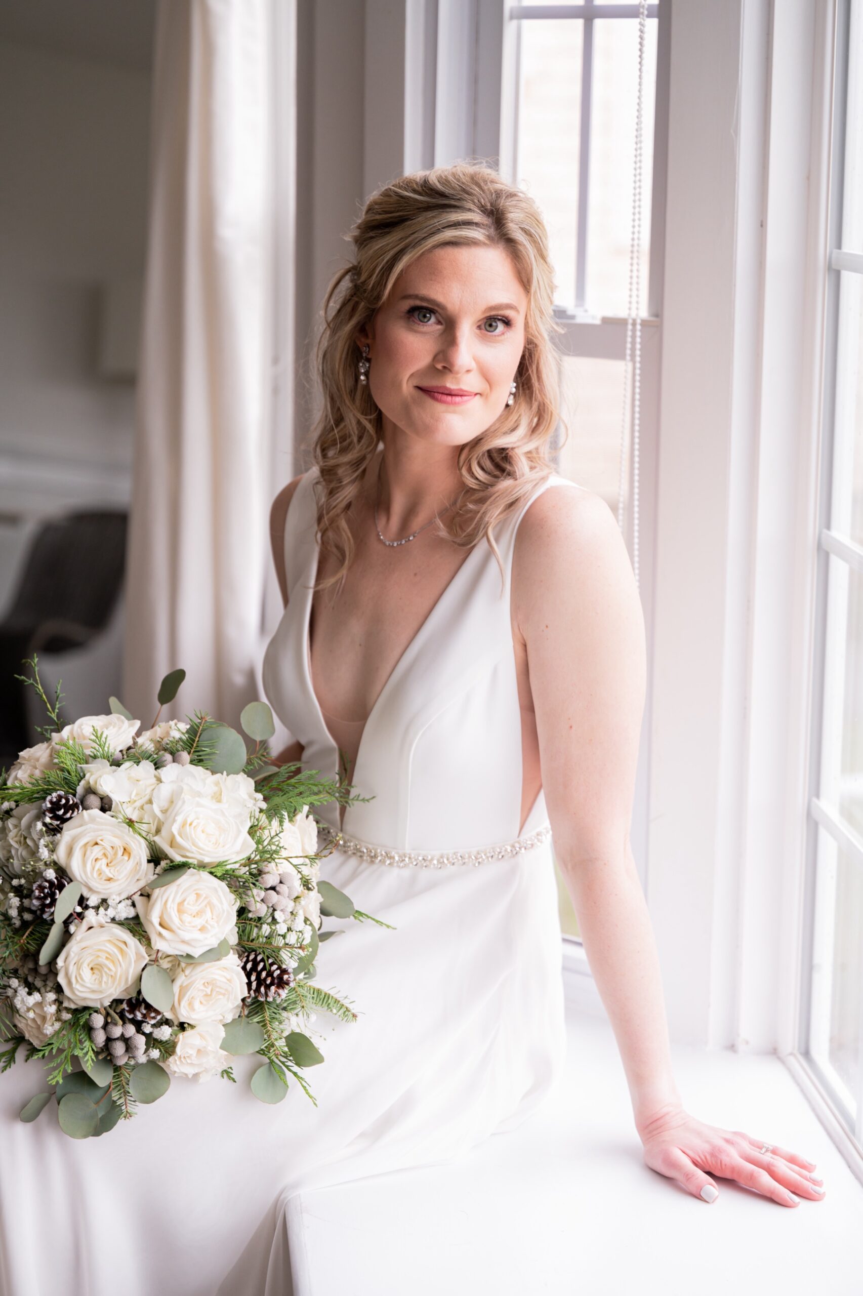 bride sitting in front of window in wedding dress and holding white and green wedding bouquet 