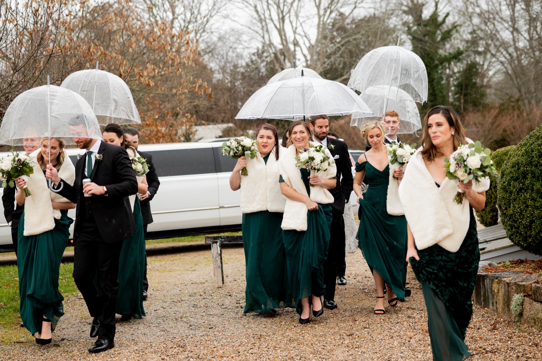 bridesmaids wearing dark green dresses and white shawls walking along the road with clear umbrellas with groomsmen