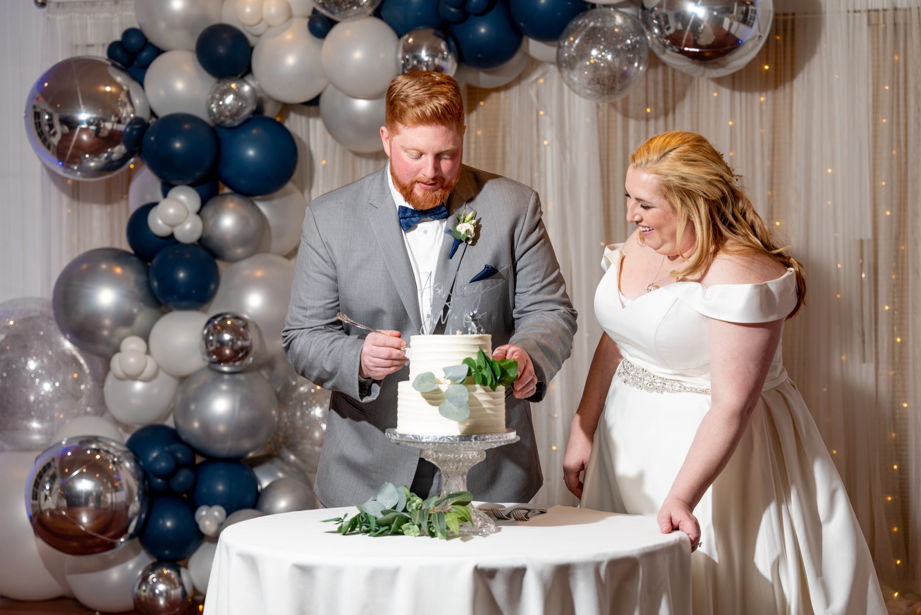 bride and groom smile as they cut their wedding cake with blue and silver balloons behind them 