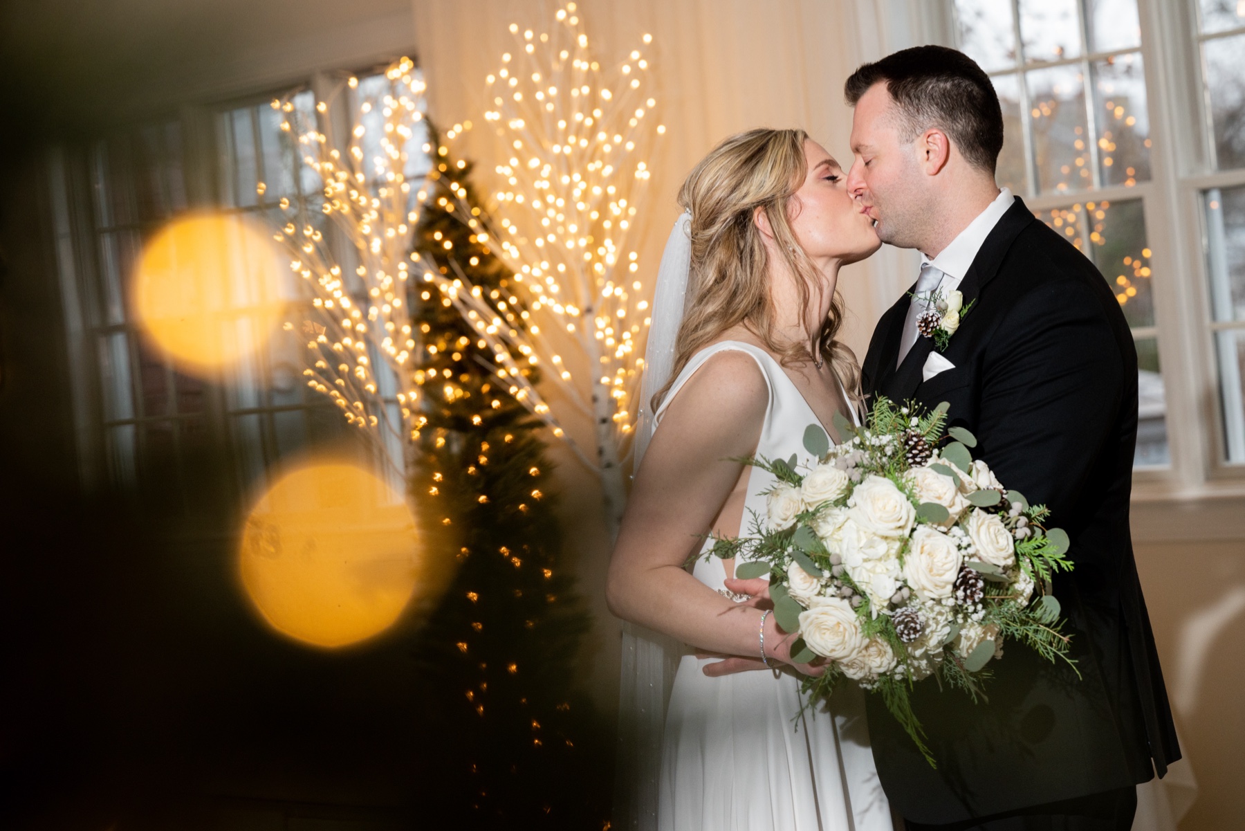 bride and groom kissing with christmas trees in the background and bride holding white and green bouquet 