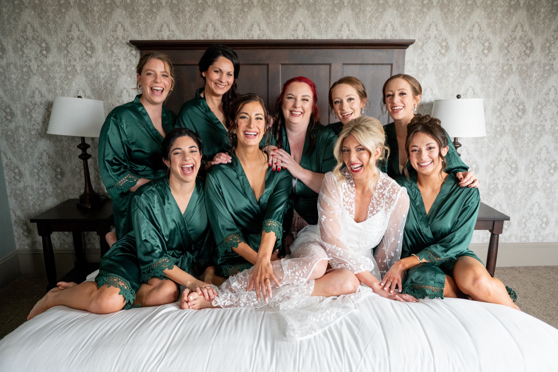 bridesmaids wearing green silk robes sitting on the bed with the bride who wears a white lace robe