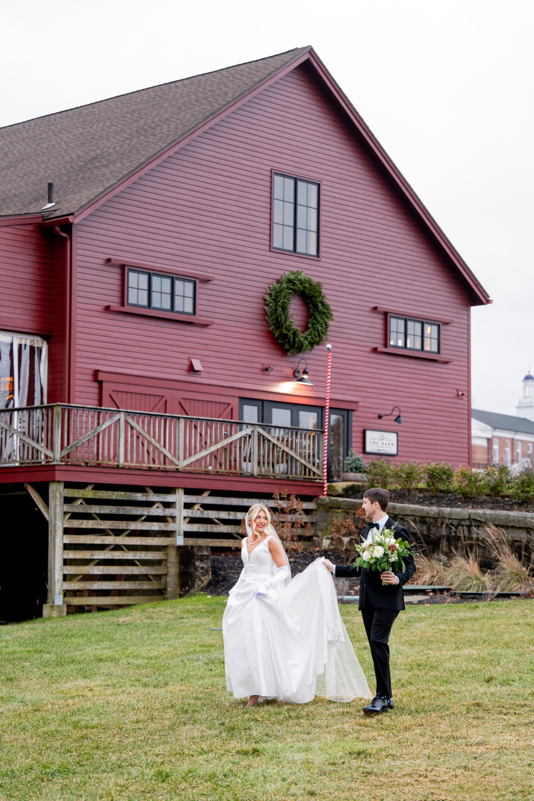 bride and groom walking in front of a red barn with a christmas wreath before their Winter Wonderland Wedding ceremony 