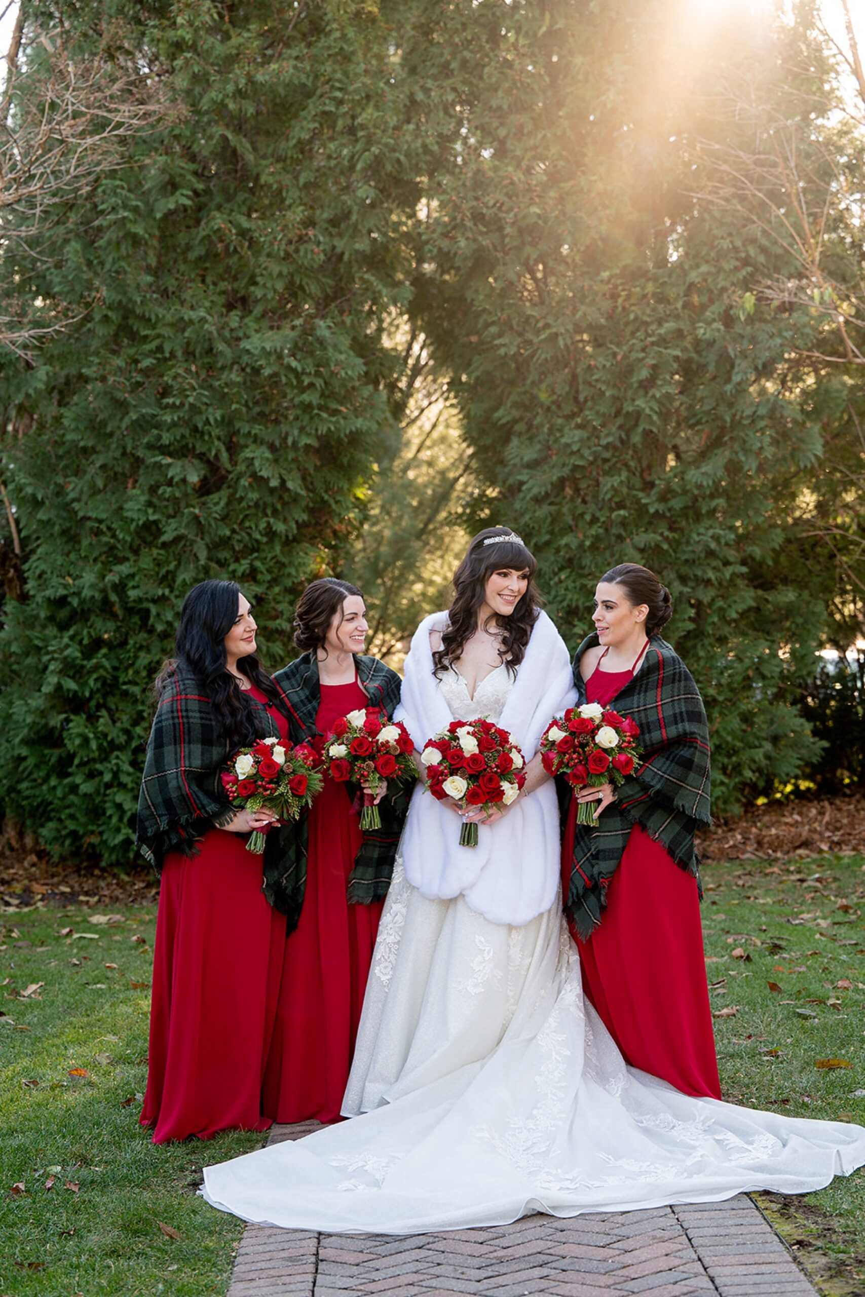 bride wearing a white fur shawl standing with her bridesmaids in red dresses and green shawls with red and white floral bouquets 