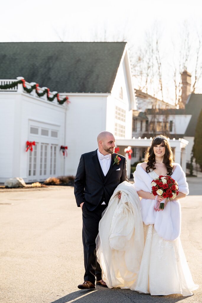 bride wearing white dress and red flower bouquet walking outside with her groom as he holds her wedding dress train 