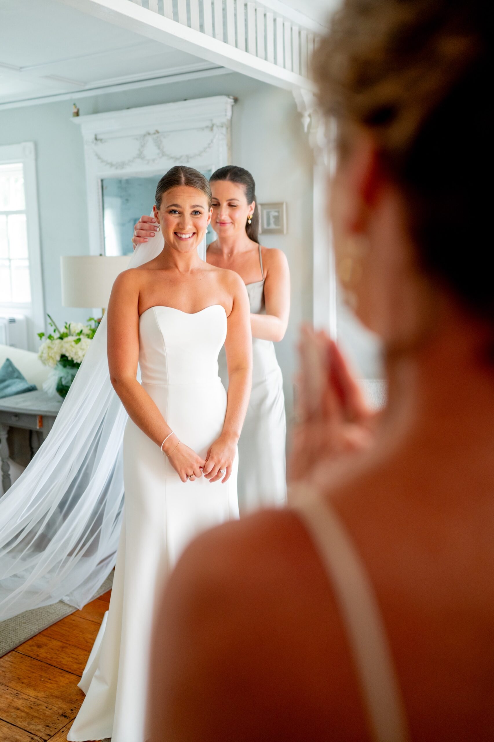 bride standing in her wedding dress smiling as her mom looks on and bridesmaid attaches her veil 