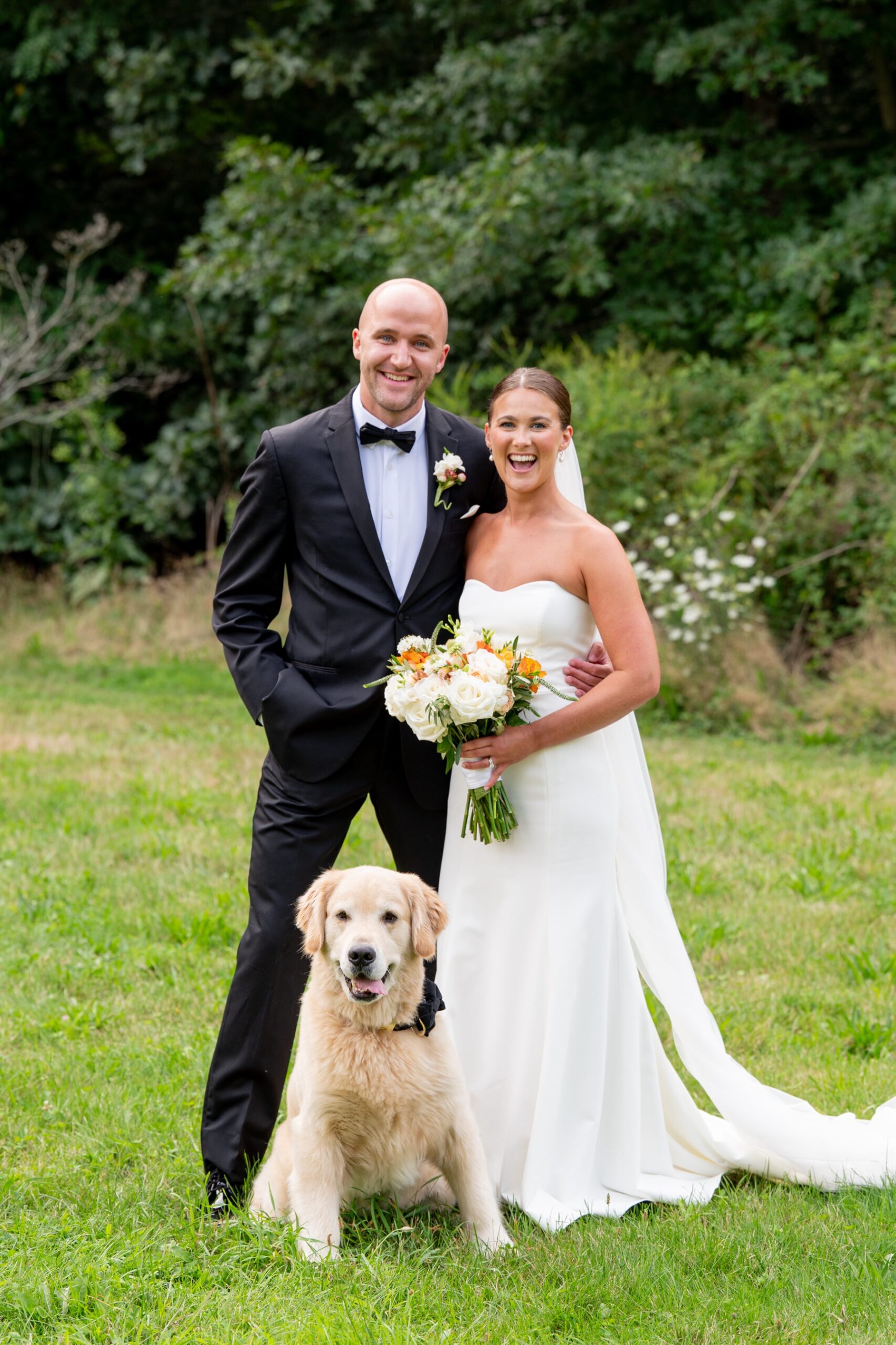 bride and groom smile at the camera as their golden retriever dog sits in front of them wearing a black bowtie during their South Shore Summer Wedding day 