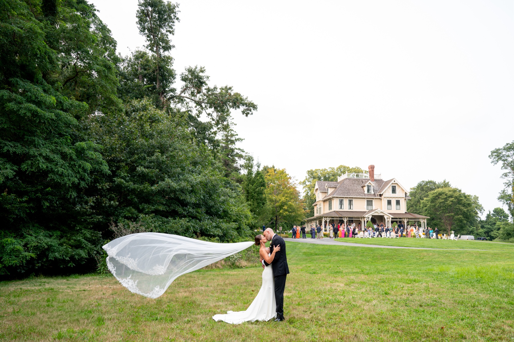 bride and groom kiss with the bride's veil flying in the wind with the cocktail hour behind them filled with guests on the front lawn of the South Shore Summer Wedding venue