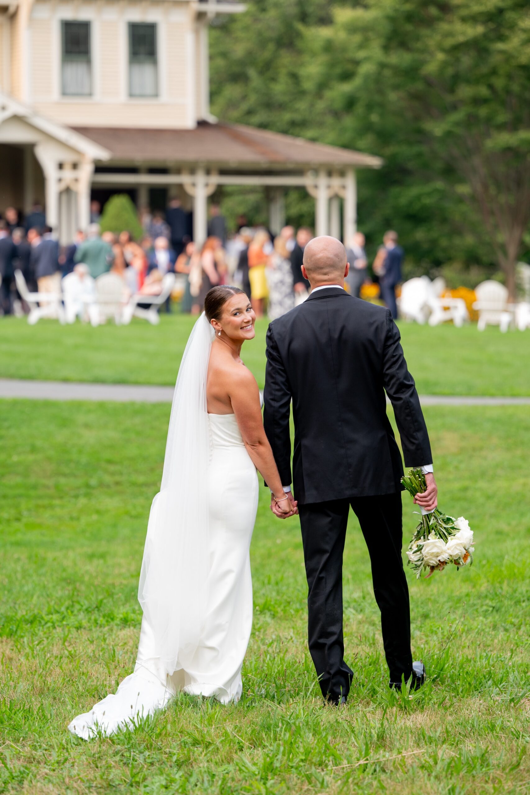 bride looking over her shoulder as she and the groom walk over to the cocktail hour on the front lawn in marshfield, MA south shore summer wedding venue