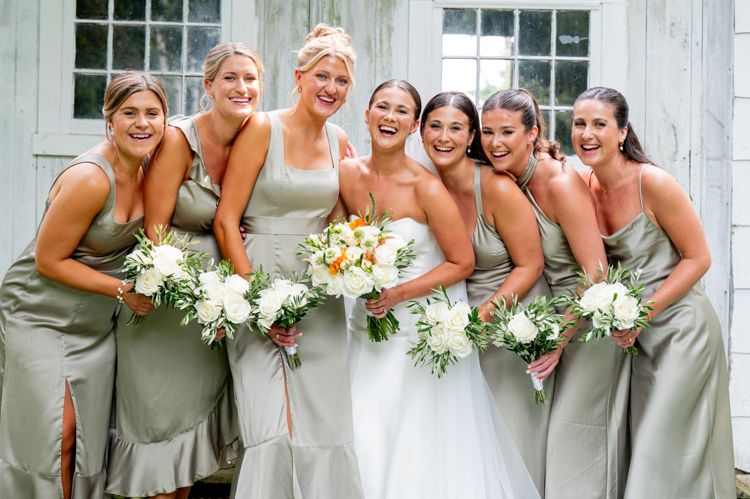 bride laughing with her bridesmaids as they stand in front of a barn on the wedding day in green dresses 