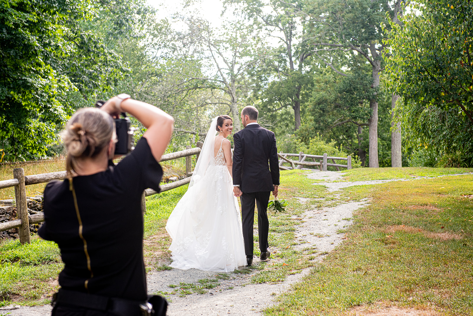 photographer taking photo of bride and groom as they walk along wooded path