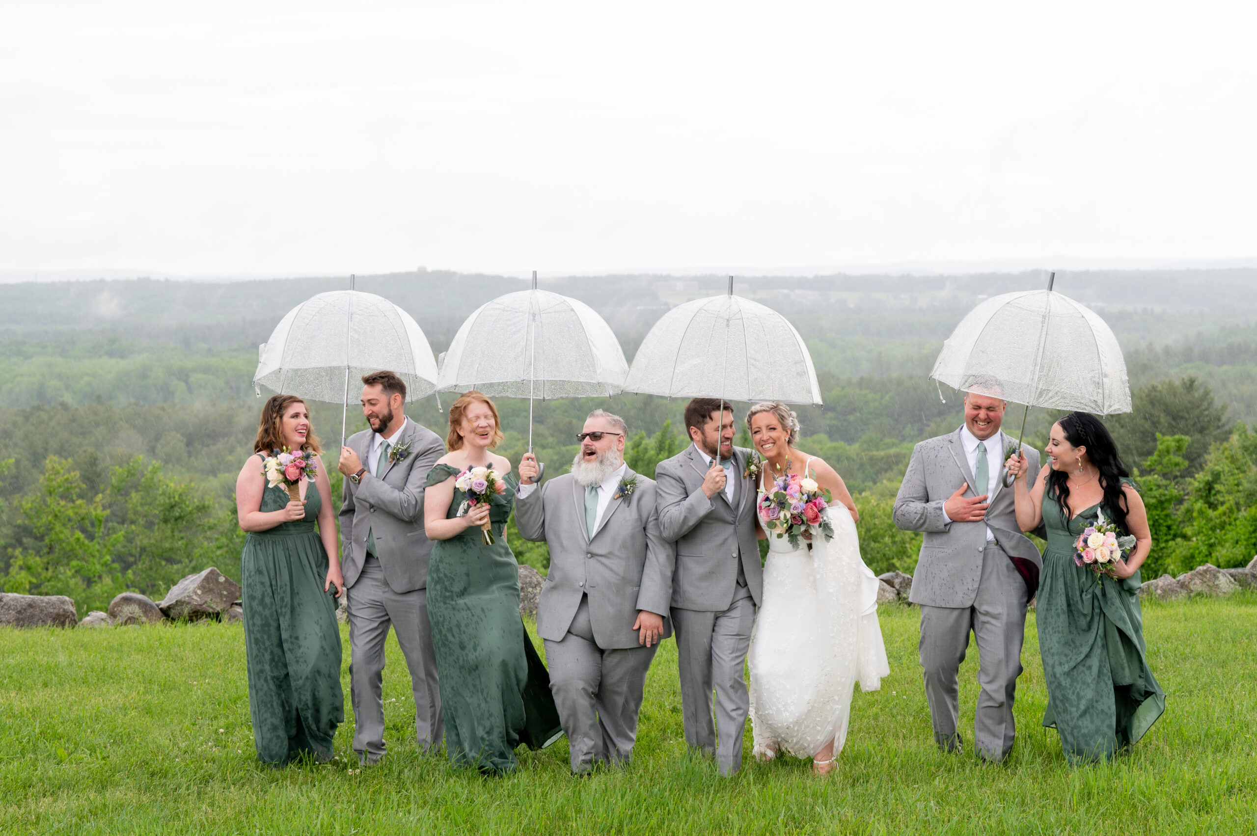 wedding party wearing green dresses and grey suits walk along a field while it's raining with clear umbrellas 
