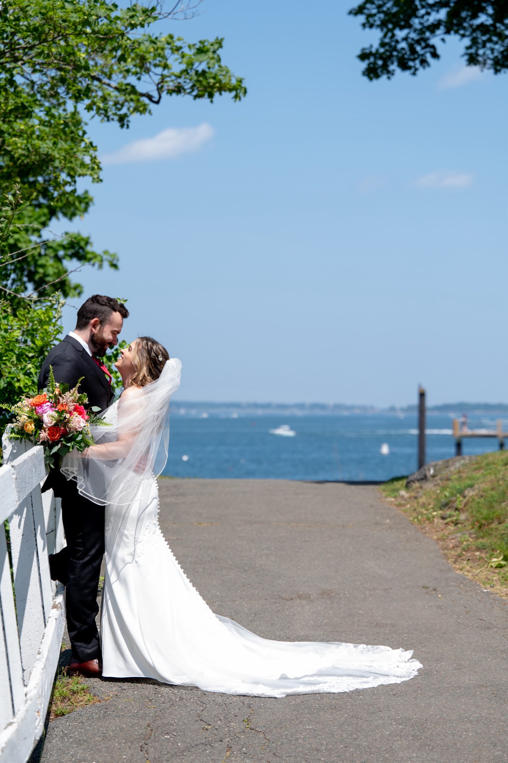 bride and groom leaning against a white fence with the ocean behind them at the Northshore of MA
