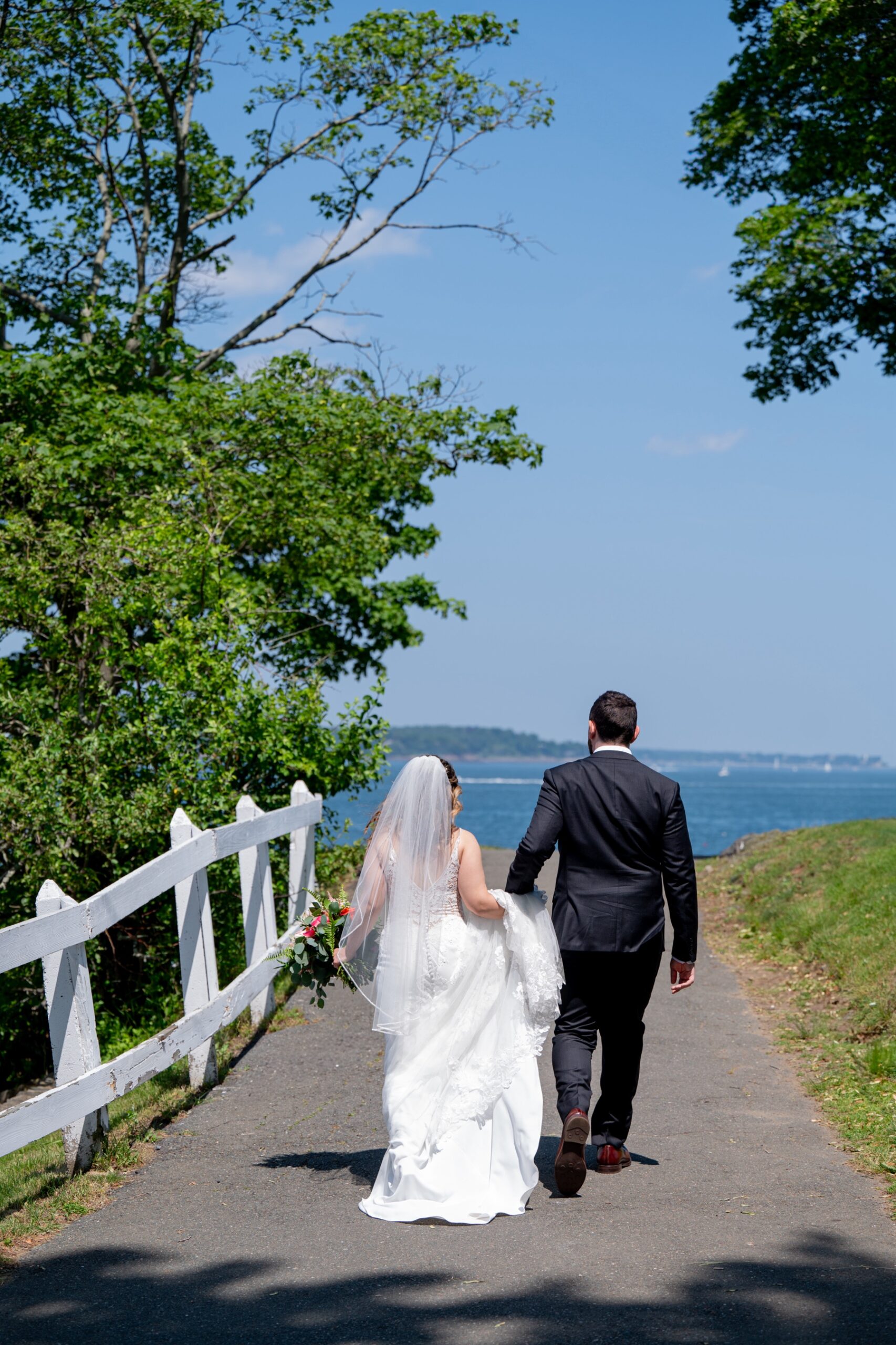 bride and groom walking down road holding hands with the ocean in front of them 