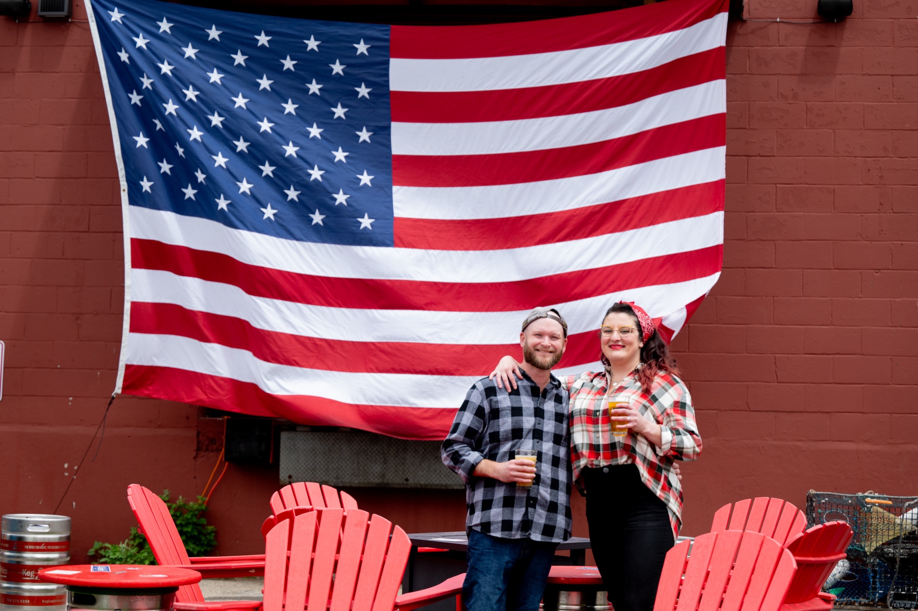 couple standing in front of big american flag holding beers 