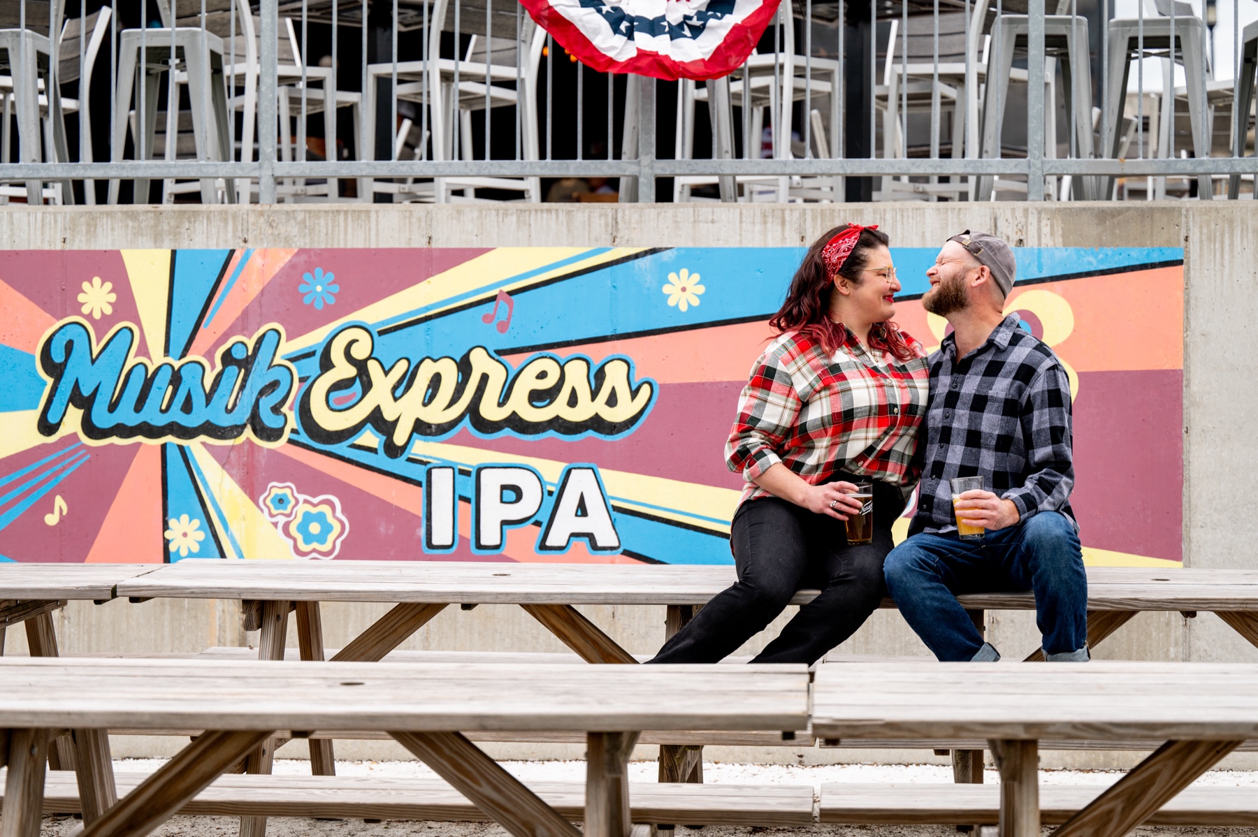 man and woman sitting on picnic table with beers in front of narragansett brewery mural