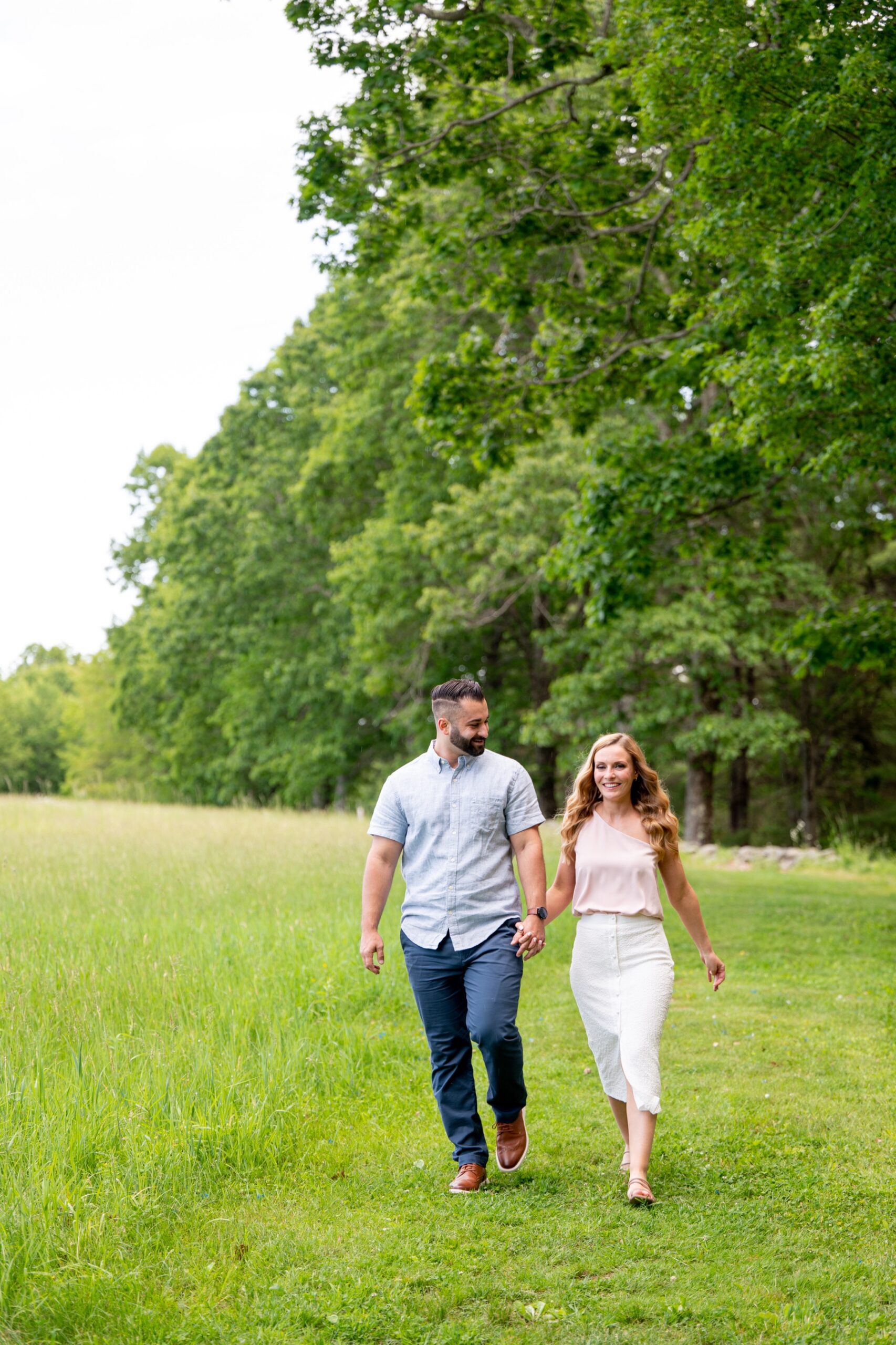 couple holding hands and walking through a green grassy field with green trees behind them